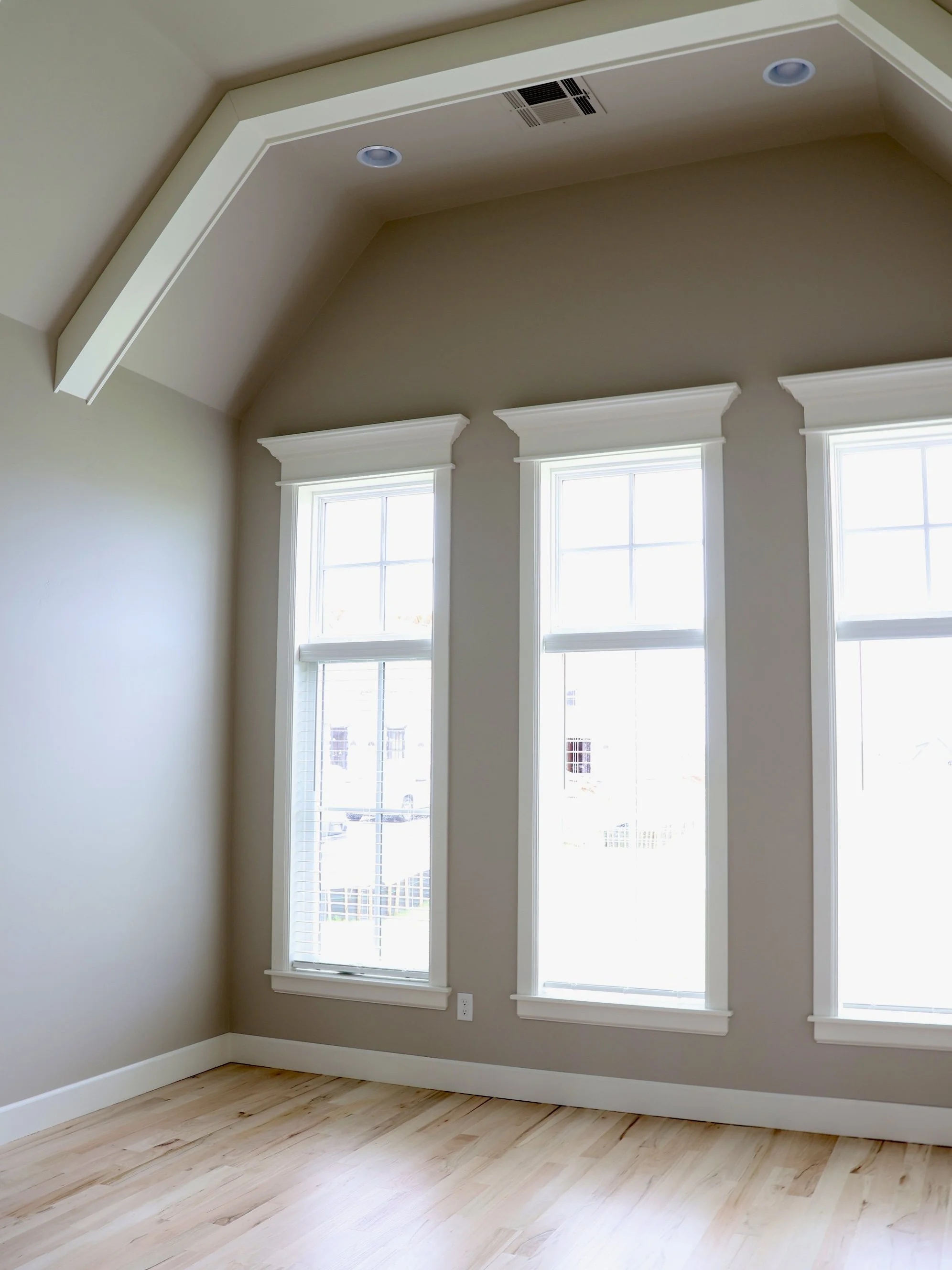 Empty room with three large windows with white trim, light-colored hardwood floors, and beige walls with white crown molding. Ceiling has two recessed lights and an air vent.