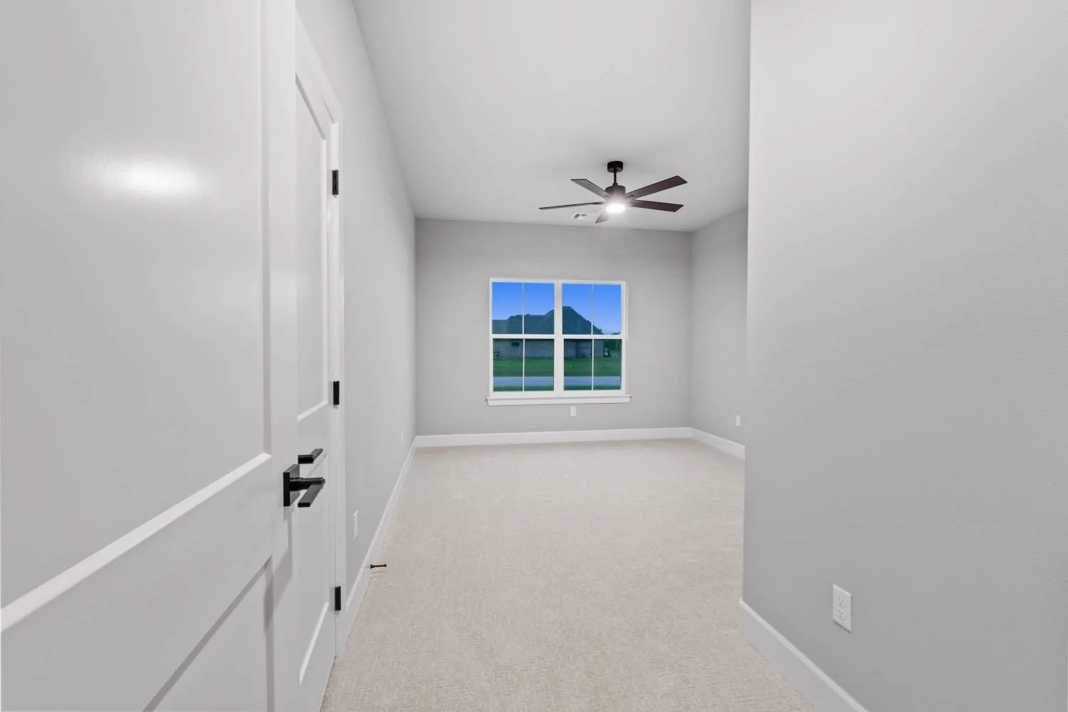 Empty room with gray walls, white baseboards, a large window showing a green lawn and house, a ceiling fan, and a closed door.