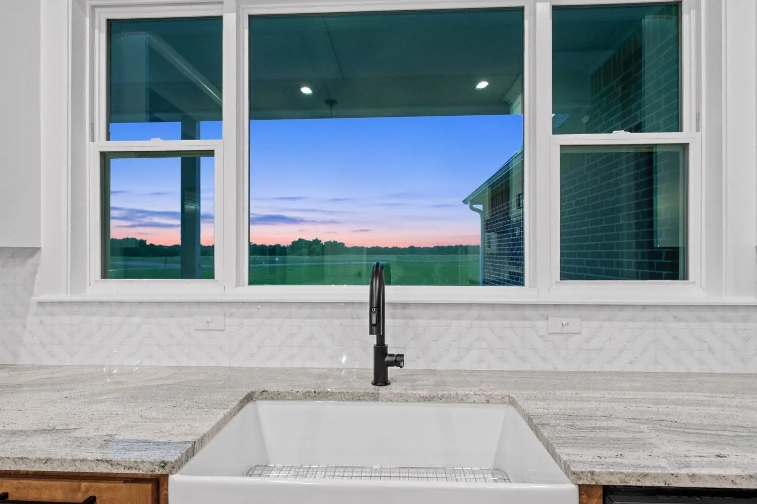 View from kitchen sink overlooking large window, showing outdoor landscape with grass, trees, and a dusk sky with clouds.