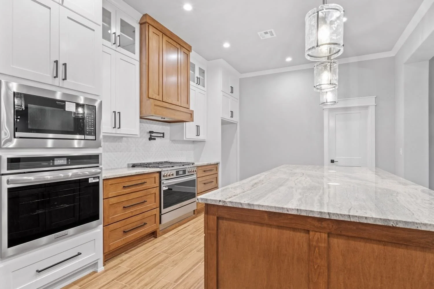 Modern kitchen with white upper cabinets, wooden lower cabinets, a marble countertop island, stainless steel oven and microwave, and decorative pendant lighting.