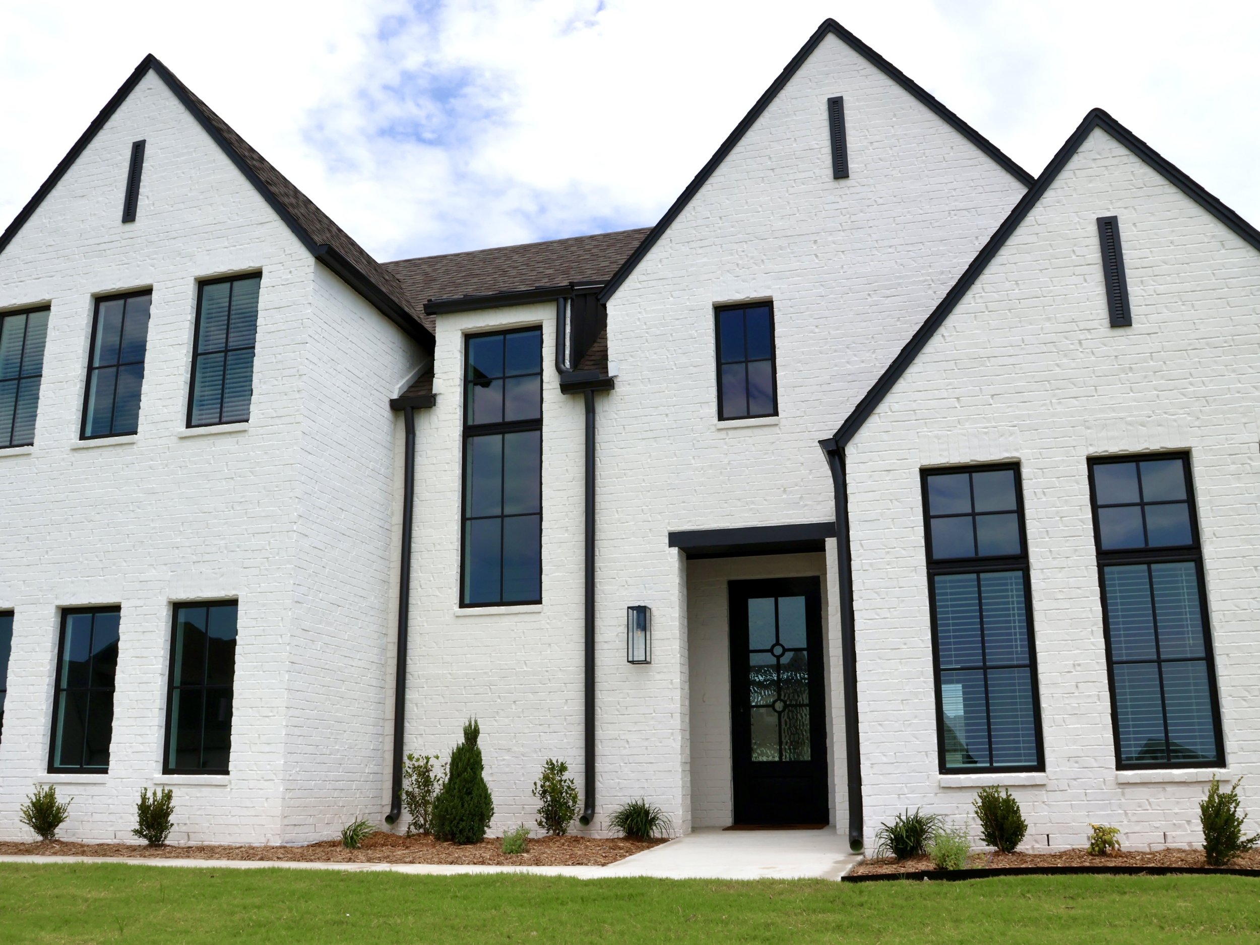 A modern white brick house with tall, pointed gable roofs and large black-framed windows. The house has a dark front door, small plants in front, and a green lawn. The sky above is partly cloudy.