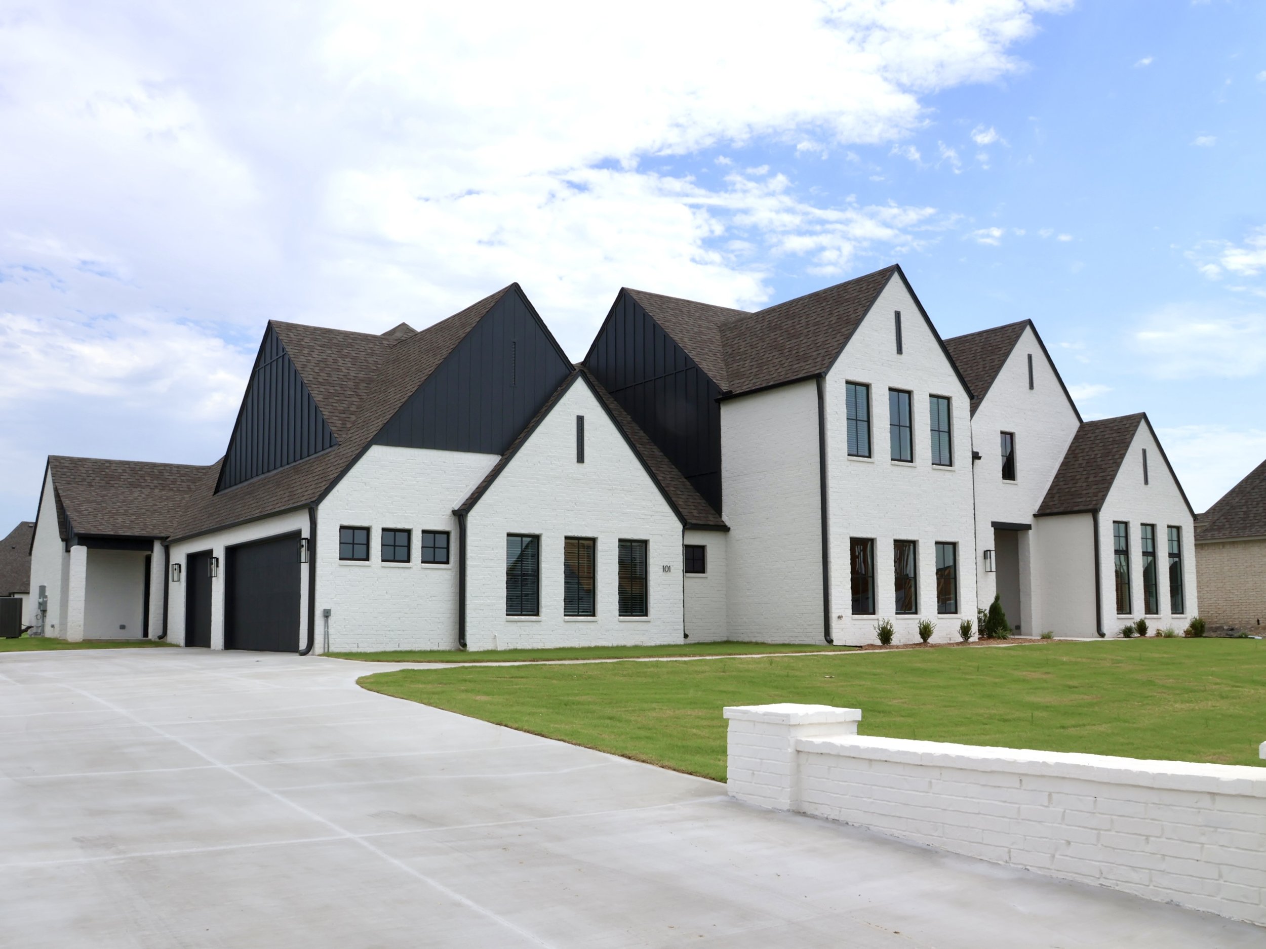 A modern white house with black and brown roof sections, multiple large windows, and a spacious driveway with a grassy lawn in front.