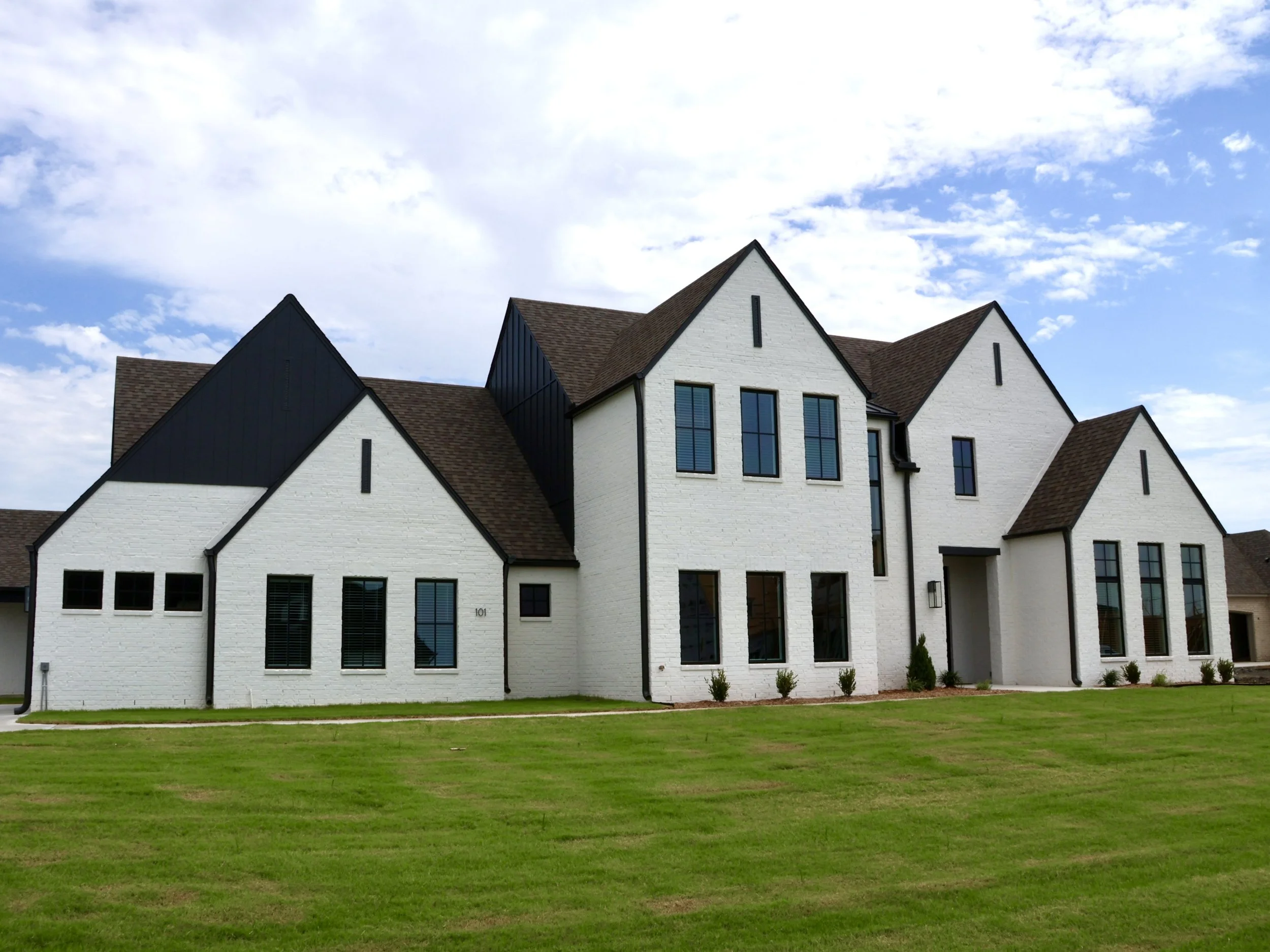 A large, modern white house with multiple steep gabled roofs, black window frames, and a well-manicured lawn in the foreground, under a partly cloudy sky.