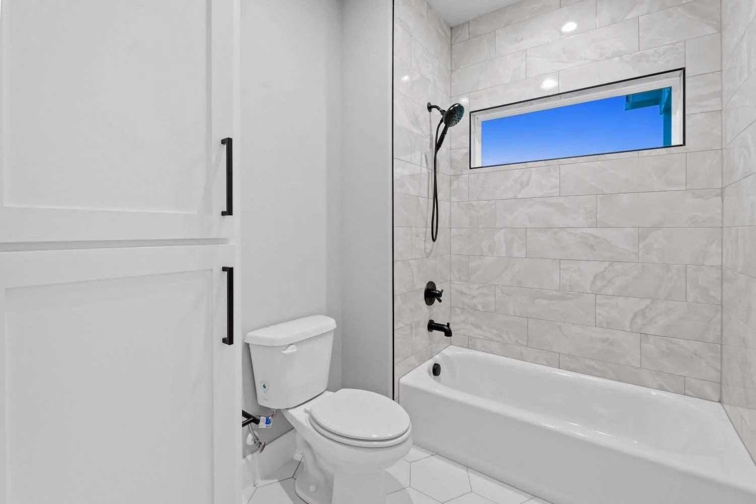 Modern bathroom with white toilet, white bathtub, gray marble tiles on wall, small horizontal window with blue sky view, and cabinetry with black handles.