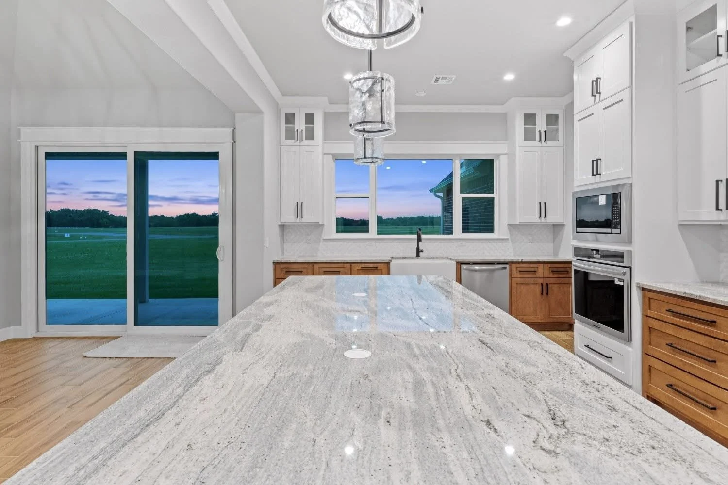 Modern kitchen with white cabinets, granite countertop island, stainless steel appliances, and large window showing a green outdoor landscape at sunset.