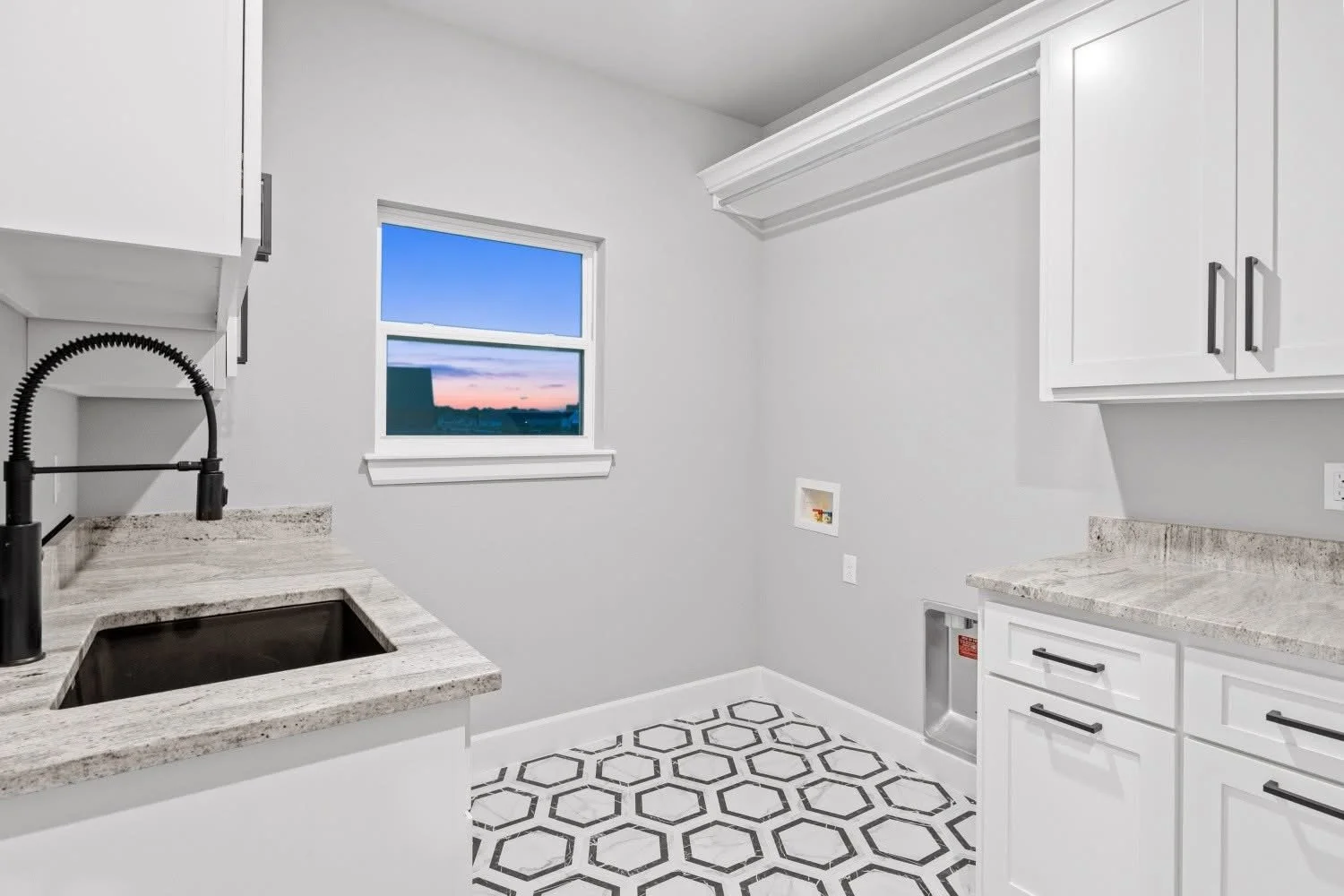 Empty modern laundry room with white cabinets, granite countertops, black faucet, hexagon patterned floor, and a window showing a sunset sky.