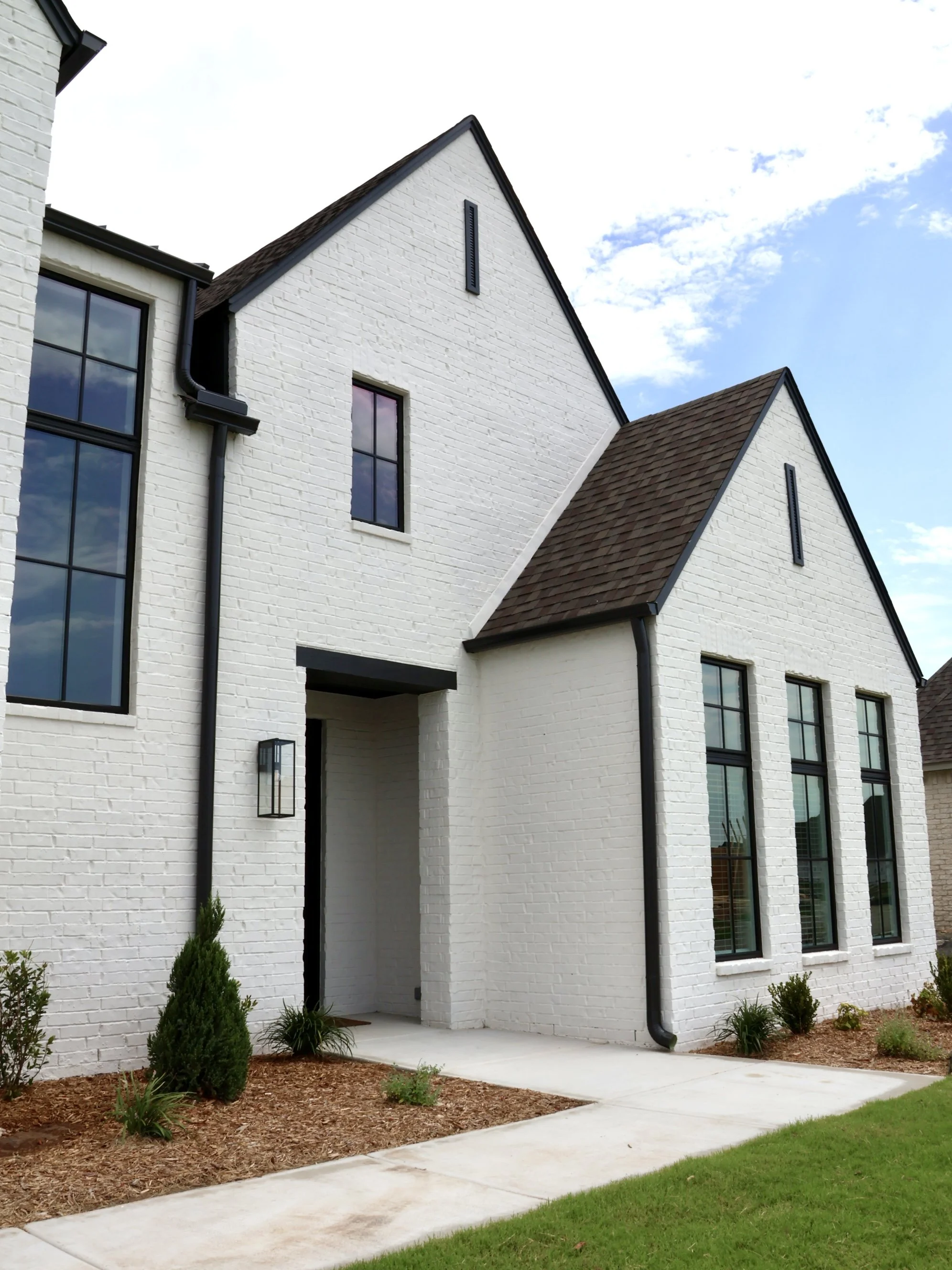 Front exterior view of a white brick house with dark roof, large windows, and landscaped yard with shrubs and pathway.