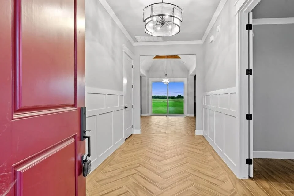Bright hallway in a house with a red front door, white wainscoting, hardwood floors, modern ceiling light, leading to a room with sliding glass doors revealing a green outdoor landscape.