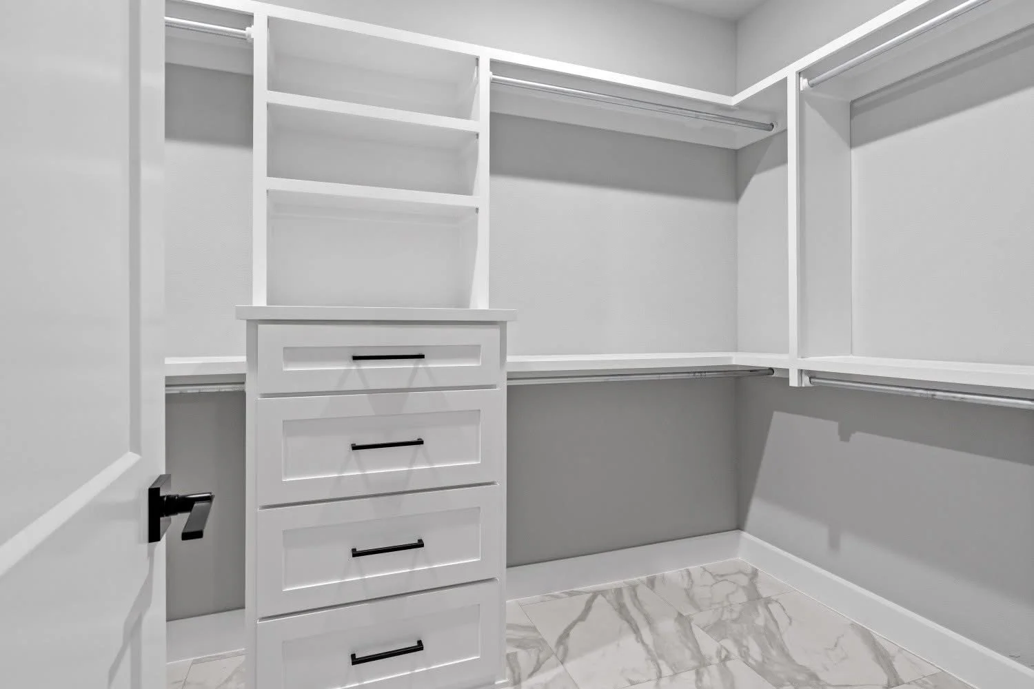 Empty white walk-in closet with built-in shelves, drawers, and hanging rods, marble flooring, and open door.