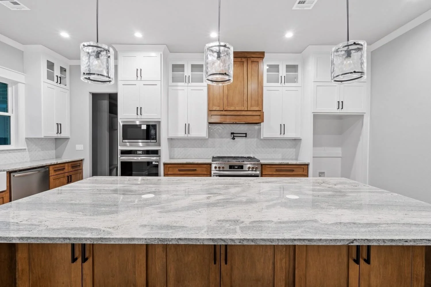 A modern kitchen with white upper cabinets, wooden lower cabinets, a large marble countertop island, stainless steel appliances, and pendant lights.