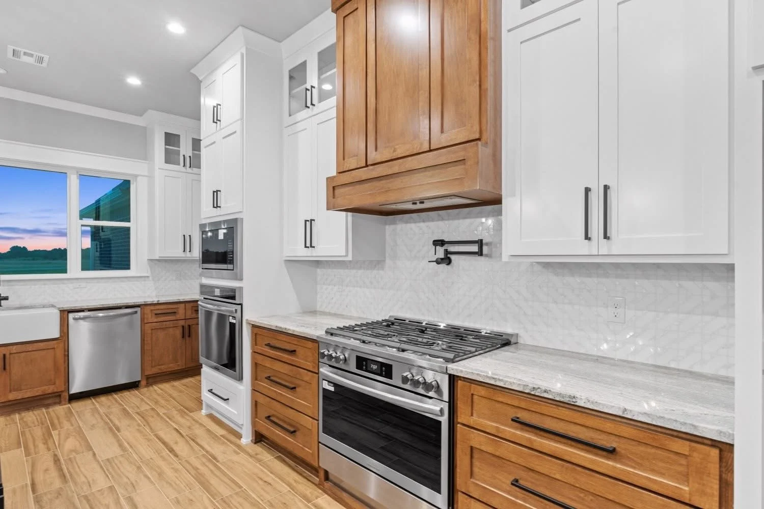 Modern kitchen with white upper cabinets, brown lower cabinets, stainless steel appliances, a granite countertop, and a window showing a sunset view.