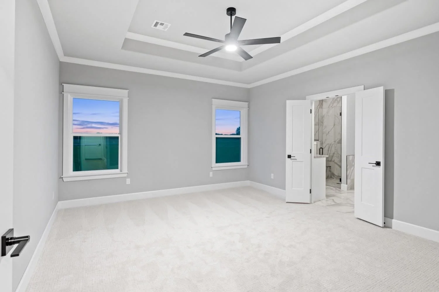 Empty bedroom with light gray walls, white trim, two windows showing a sunset sky, a ceiling fan, and an open door leading to a marble-tiled bathroom.