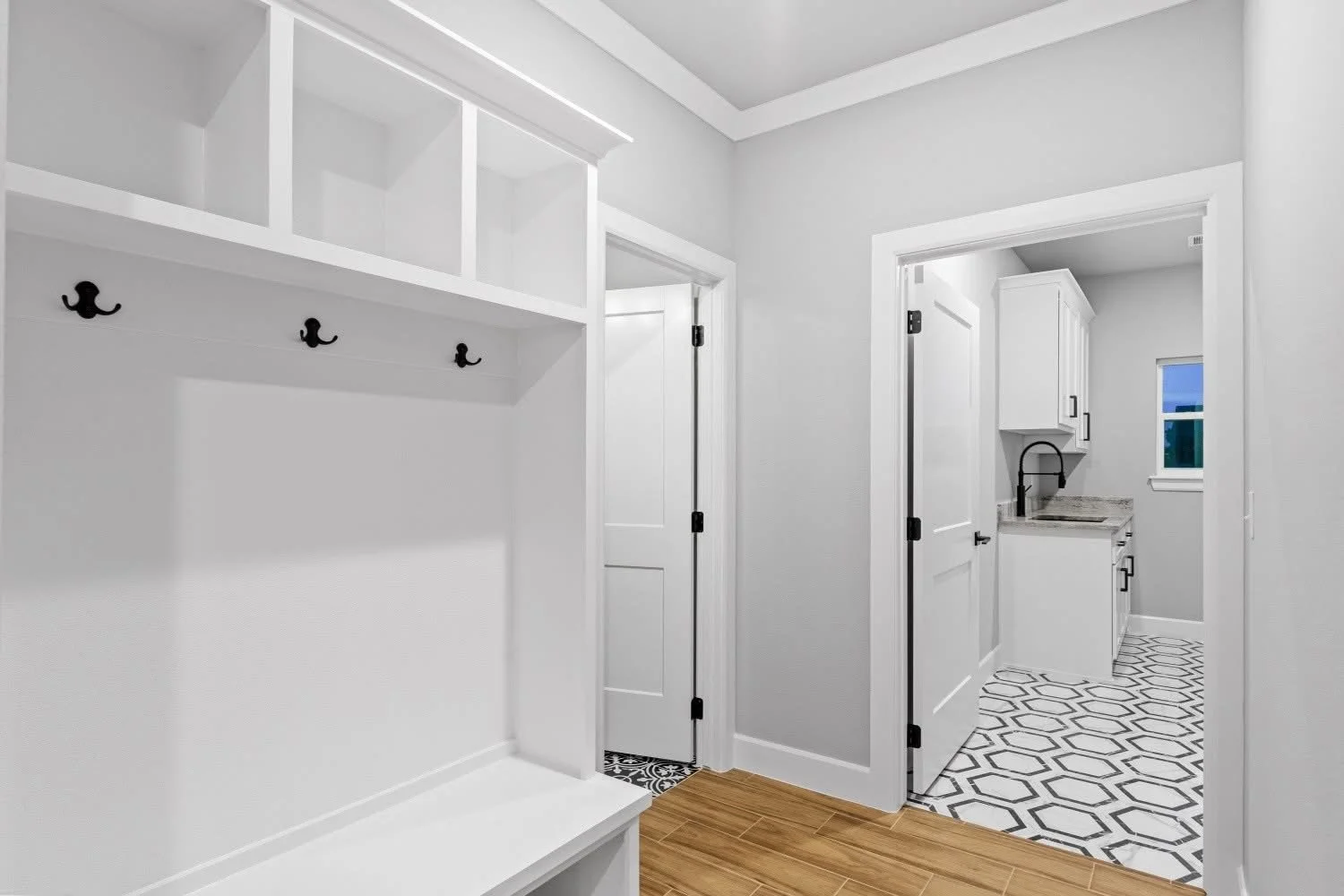 A bright, modern laundry or mudroom with white built-in cabinetry, black hooks on the wall, wood flooring, and a small utility sink area in a connected room with patterned tile flooring.