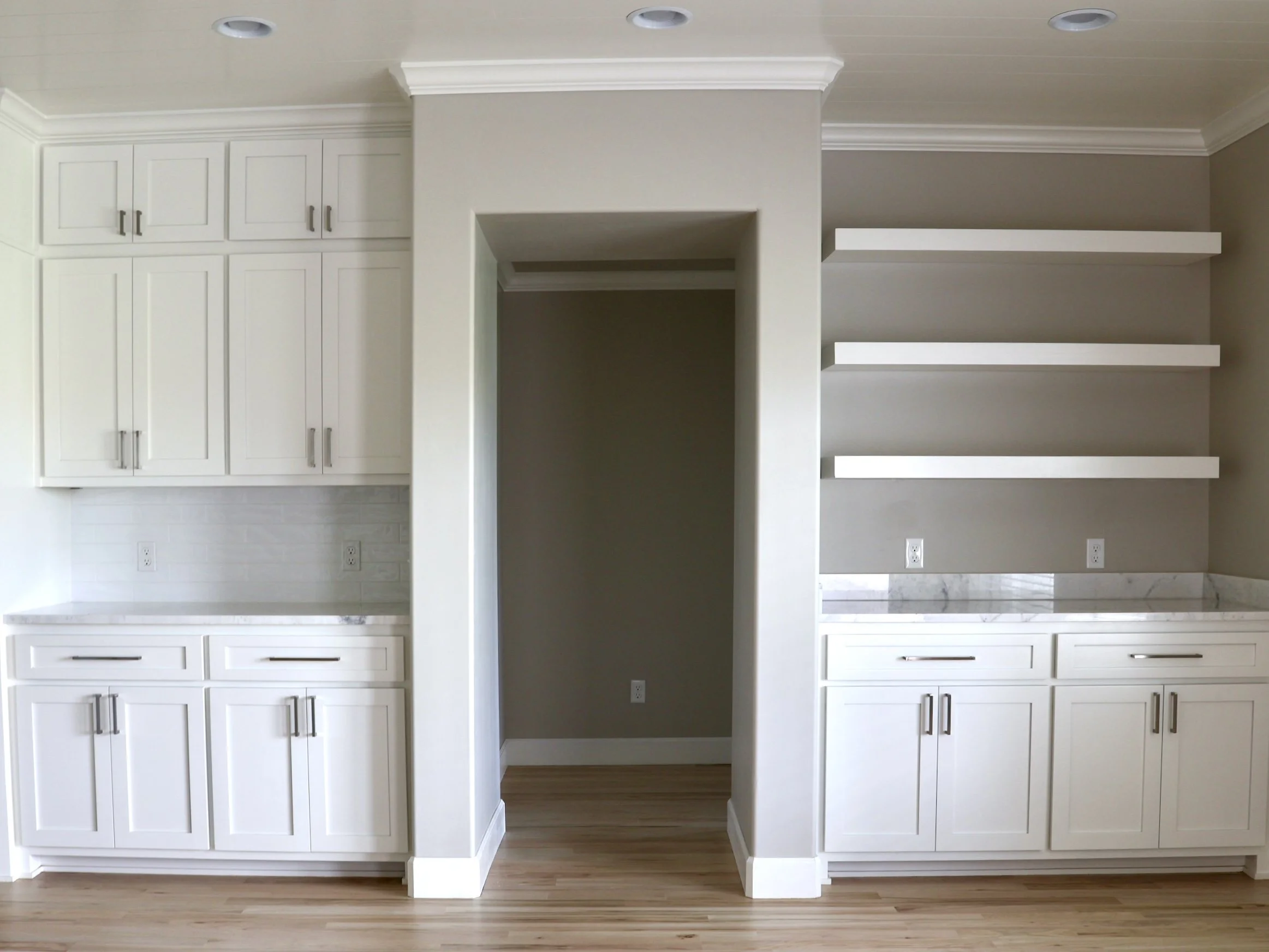 A white built-in cabinet with closed cabinet doors on either side, three open shelves on the right, and partial white cabinetry below, against a wall with electrical outlets and a wooden floor.