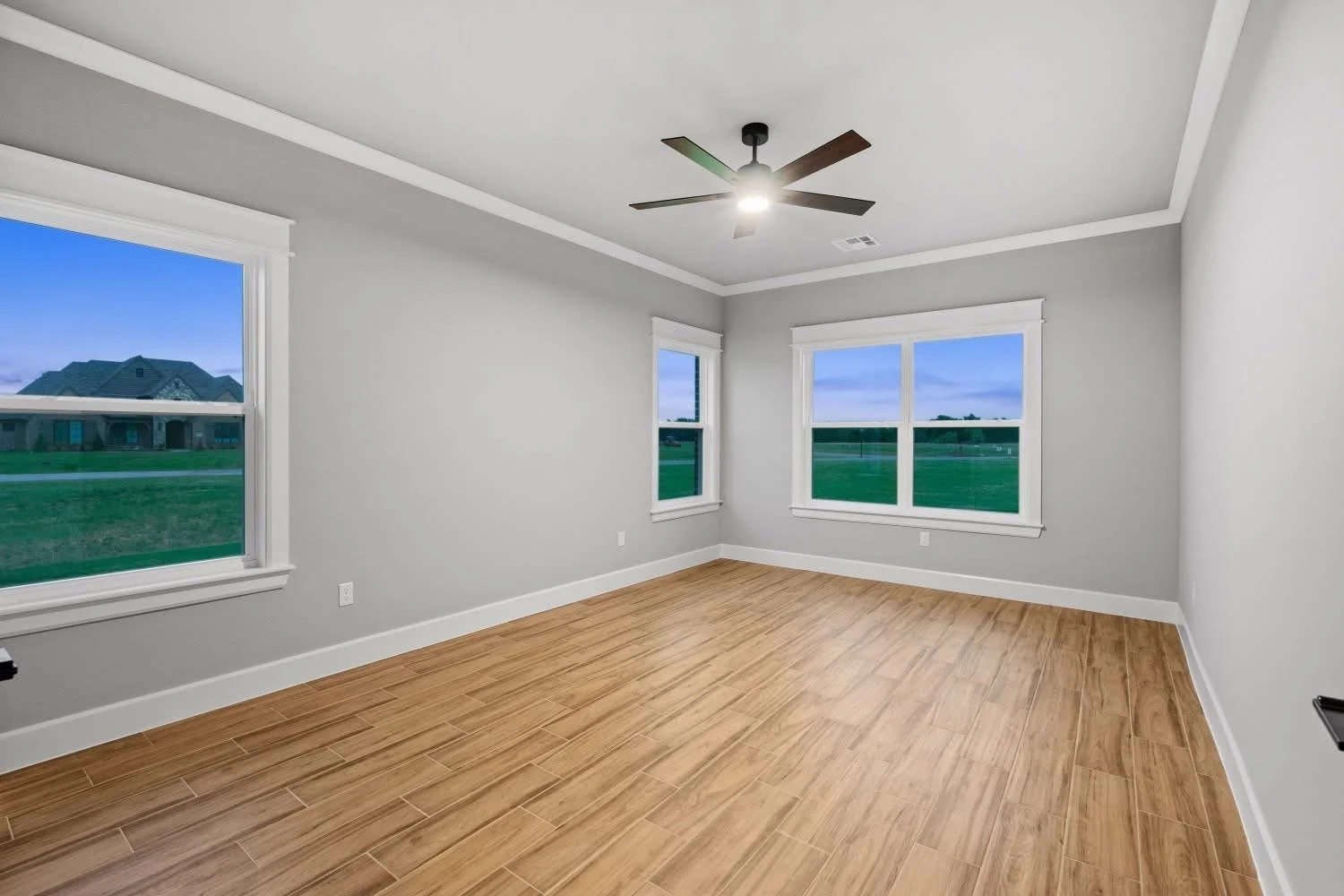 Empty modern room with light gray walls, white trim, three large windows showing a green field outside, a wood floor, and a ceiling fan with lights.