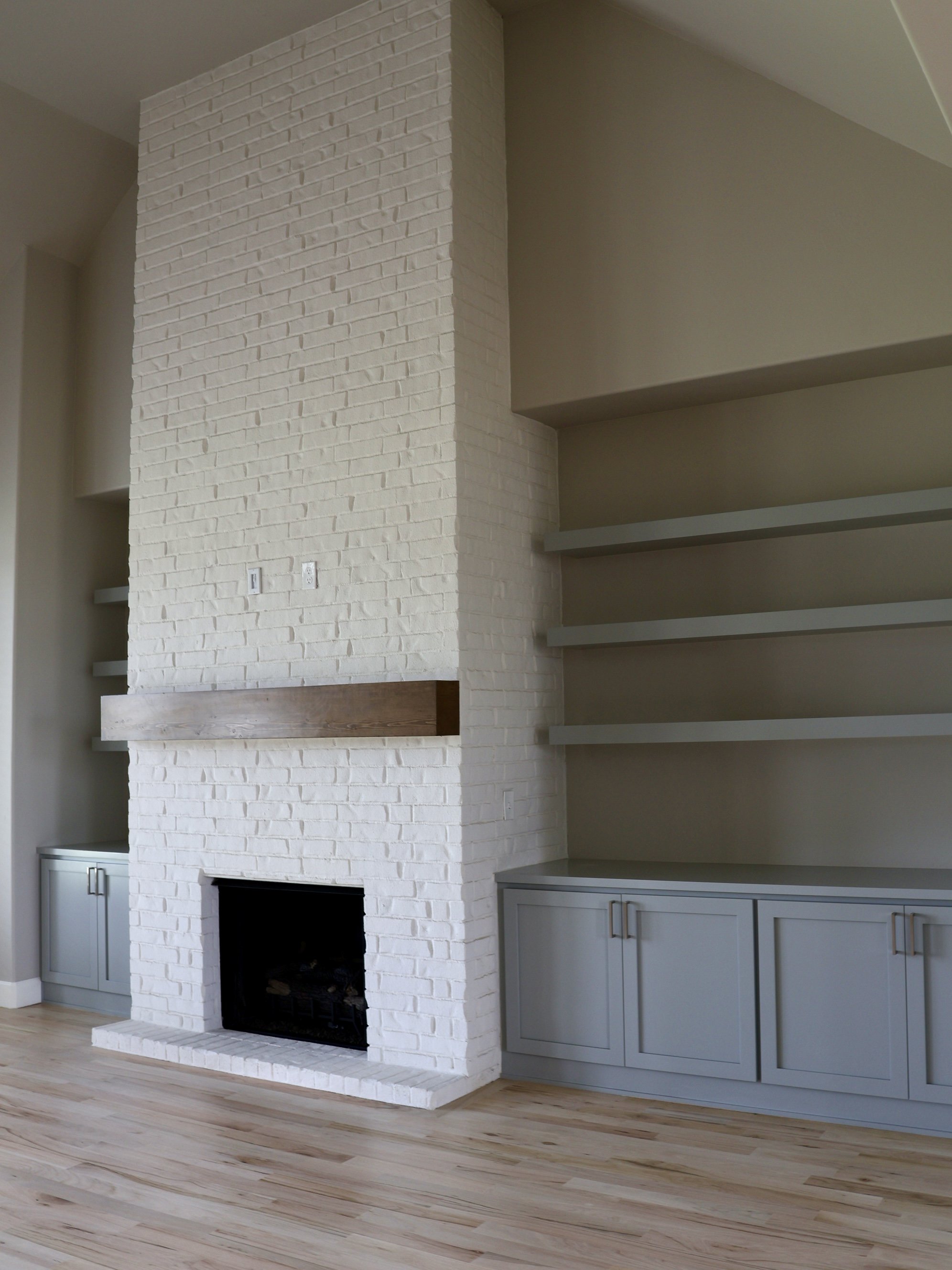 Interior of a living room with a white brick fireplace, gray built-in cabinets, and open shelves, with light wood flooring.