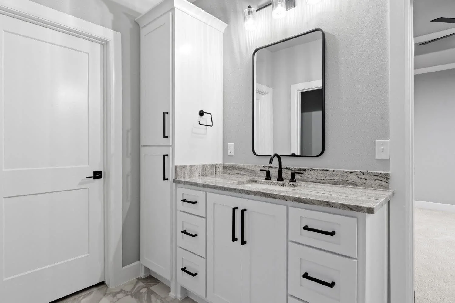 White bathroom vanity with black handles, granite countertop, black faucet, wall mirror, gray walls, and a door to the left.