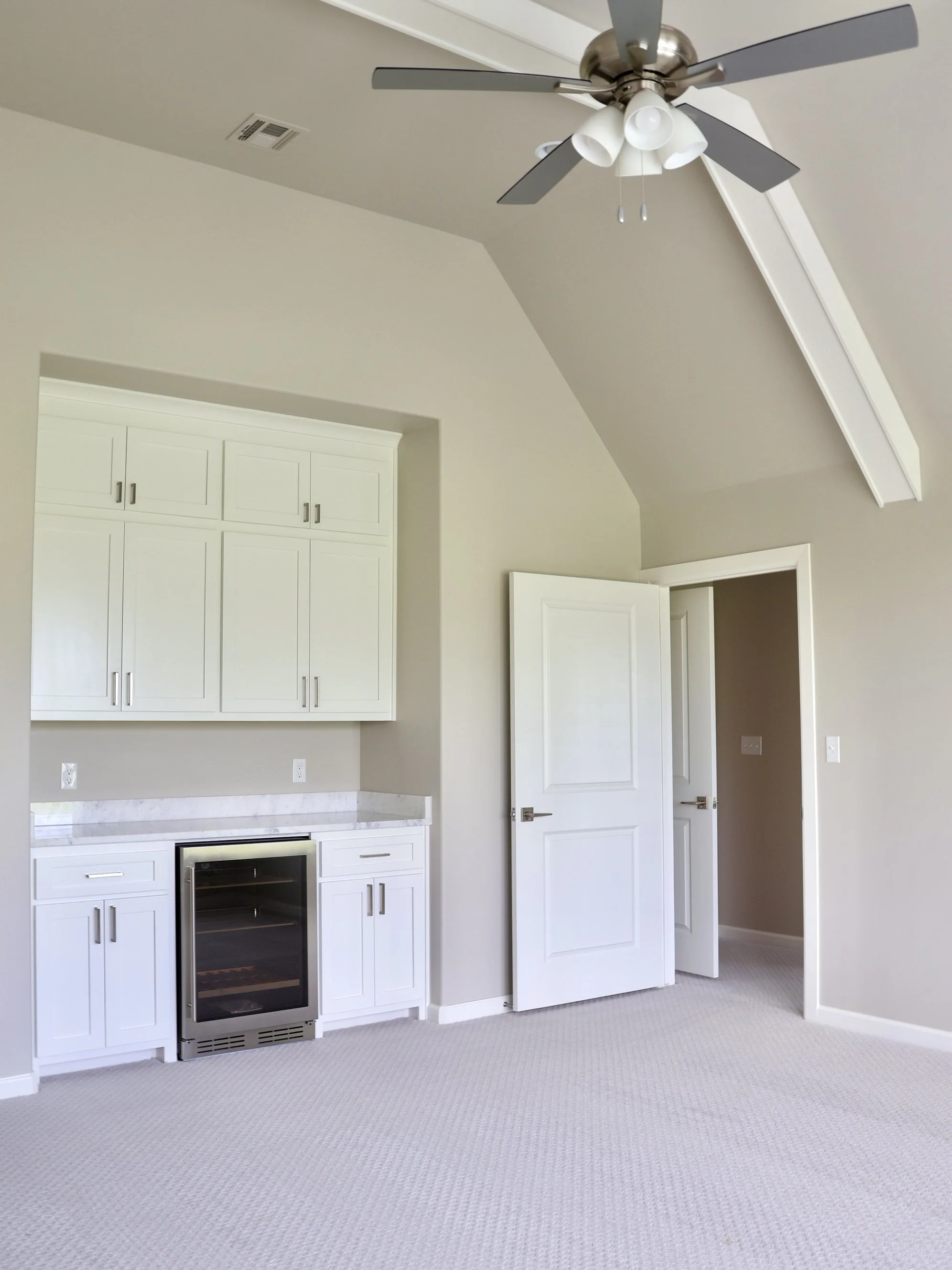 Interior view of a room with a white kitchen cabinet, a small wine cooler, and a ceiling fan with light fixtures, white walls, and beige carpeted floor.