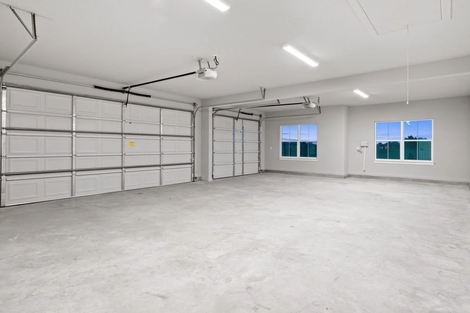 Empty two-car garage with white walls, concrete floor, and two windows showing an outdoor view, with garage doors and ceiling lights.