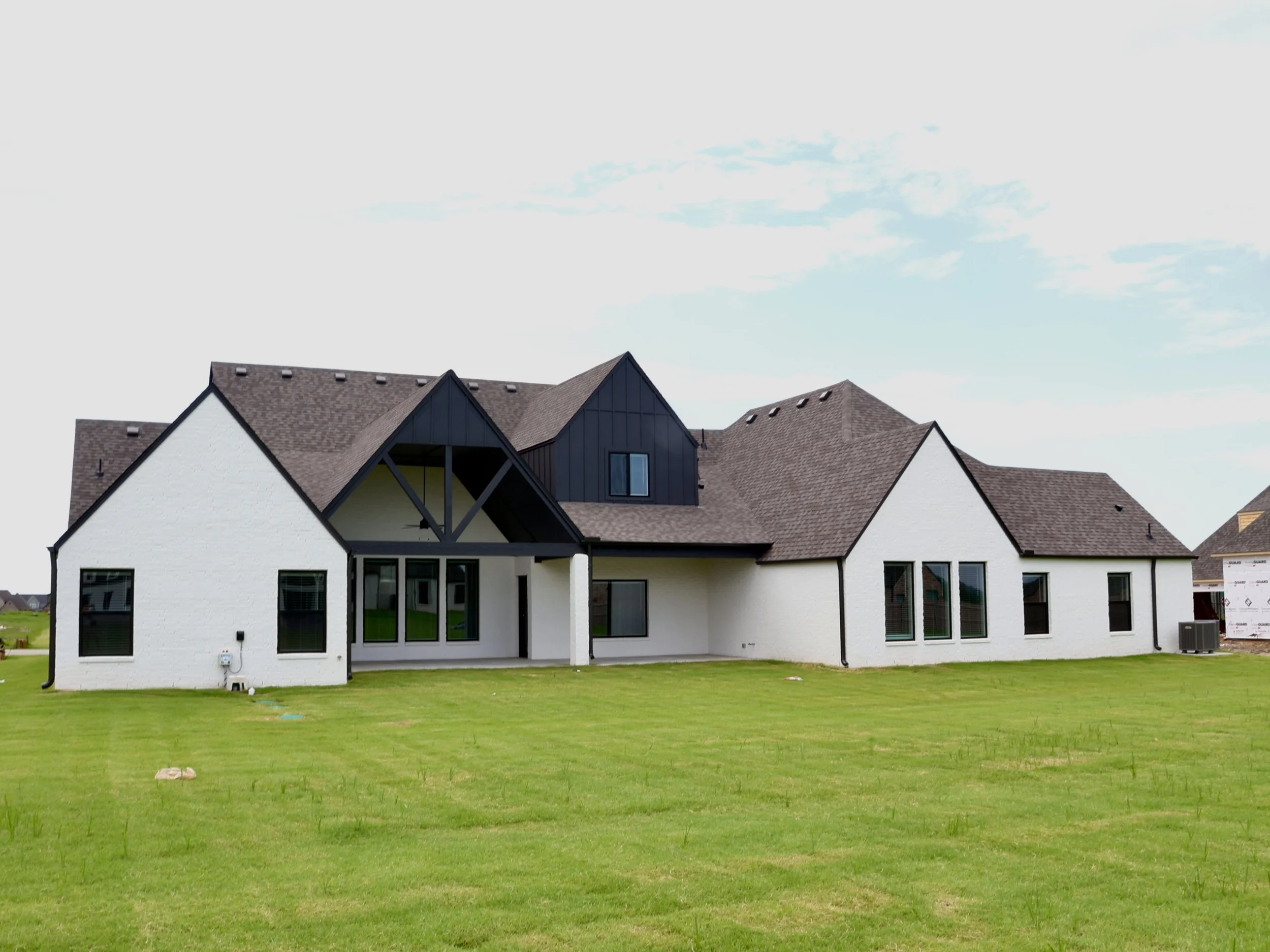 A house with a modern design, white exterior walls, black accents, and a complex roof, situated on a green lawn under a partly cloudy sky.