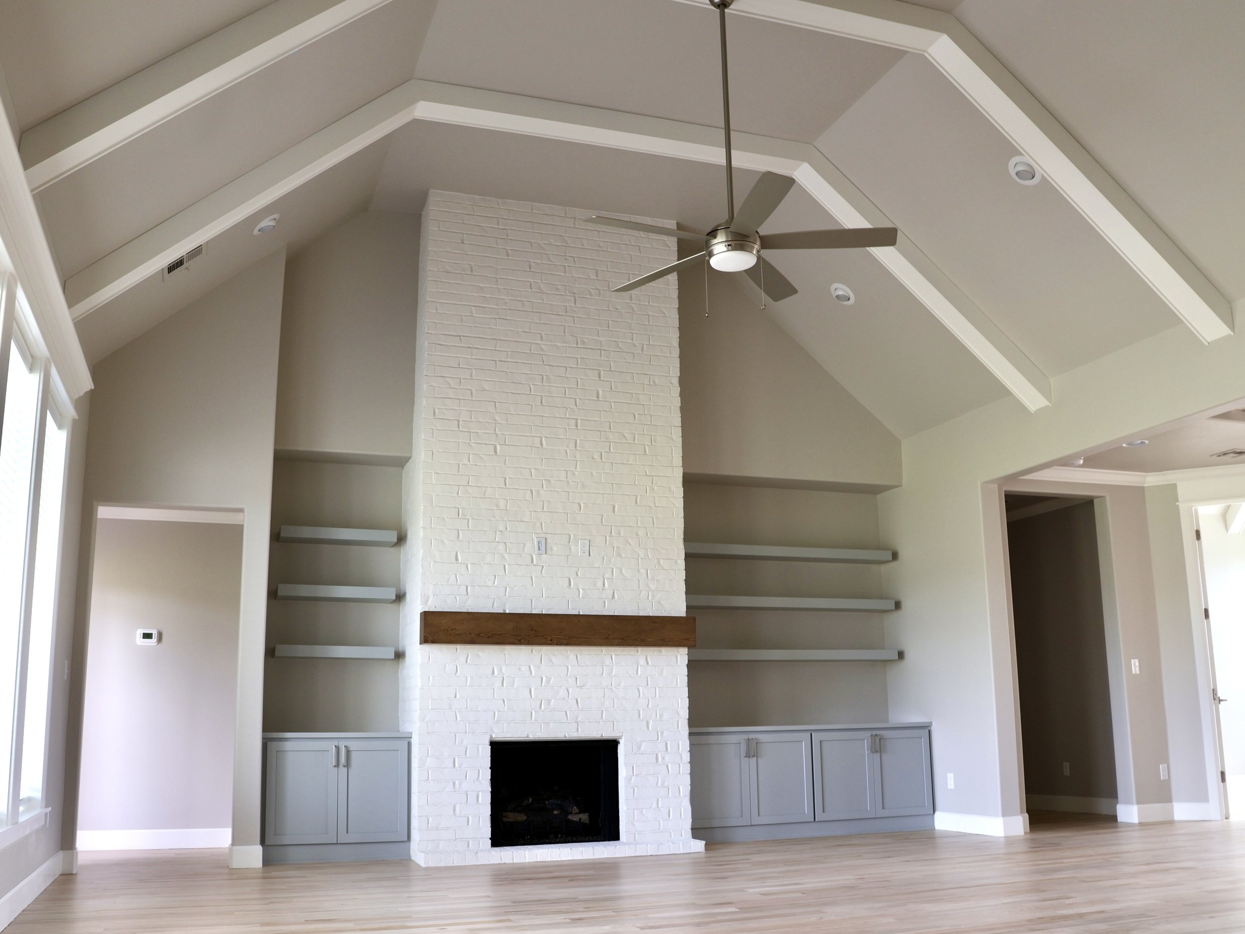 Living room with a white brick fireplace, built-in gray cabinets, and open shelving above. A ceiling fan with a light is mounted on the vaulted ceiling. Light wood flooring and a large window are visible.