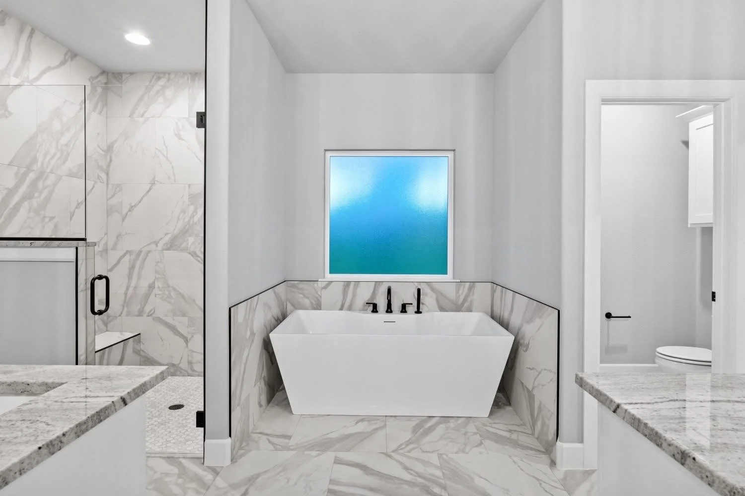 Modern bathroom featuring a freestanding bathtub with black fixtures, large marble tiles, a frosted window, a glass-enclosed shower, and a separate toilet room.