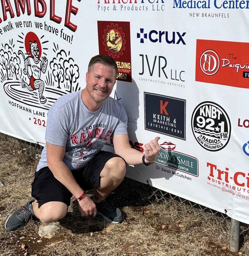 James Keith outdoors, smiling, and pointing at a large sponsorship banner with various company logos, at an event called Hoffman Lane 2022.