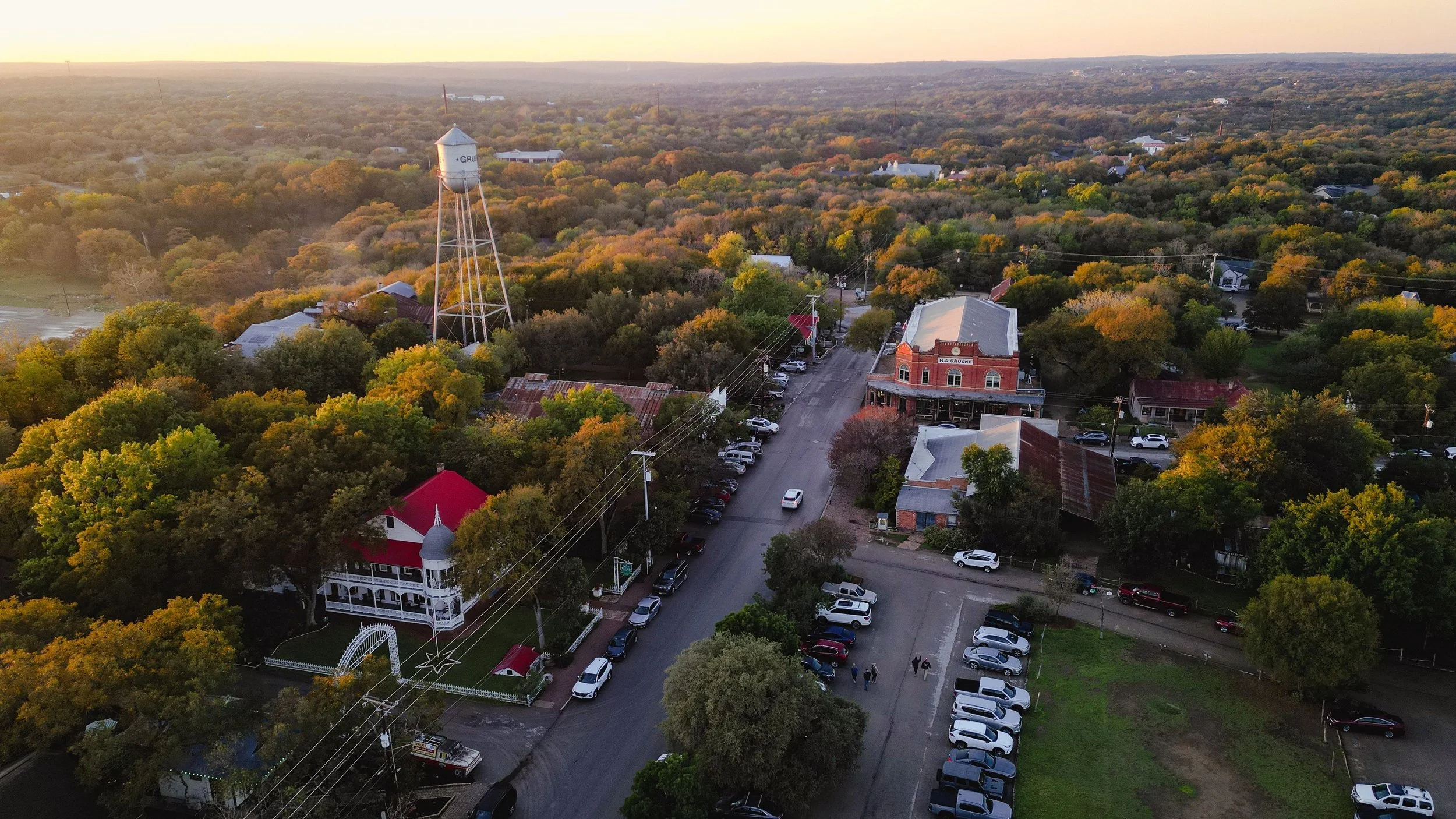 Aerial view of New Braunfels, Texas with trees, buildings, and cars during sunset. A water tower and a historic brick building are visible among the greenery.