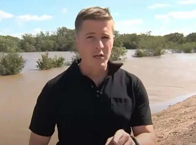 James Keith reporting on TV standing near a body of water, with trees and shrubs in the background, during daytime.