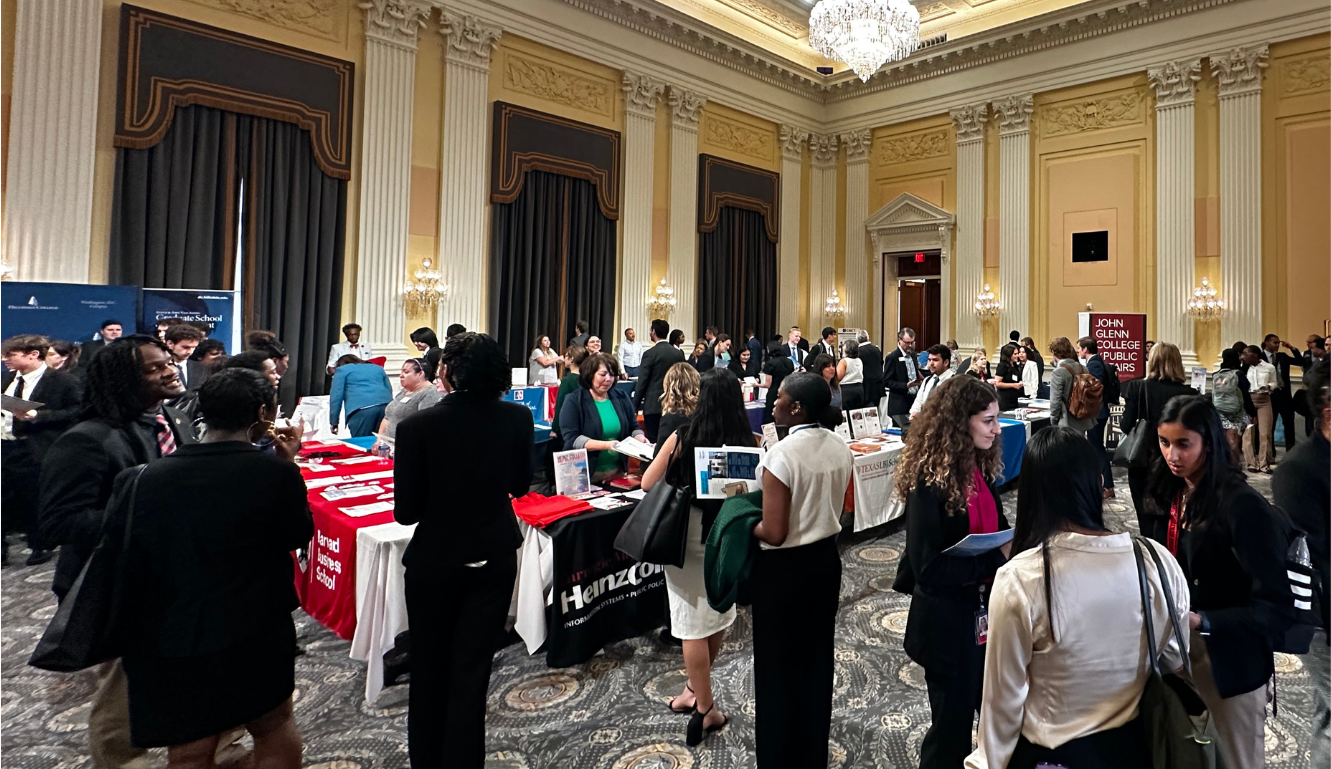 A busy grad fair with numerous booths and attendees inside an ornate ballroom with high ceilings, chandeliers, and gold accents.