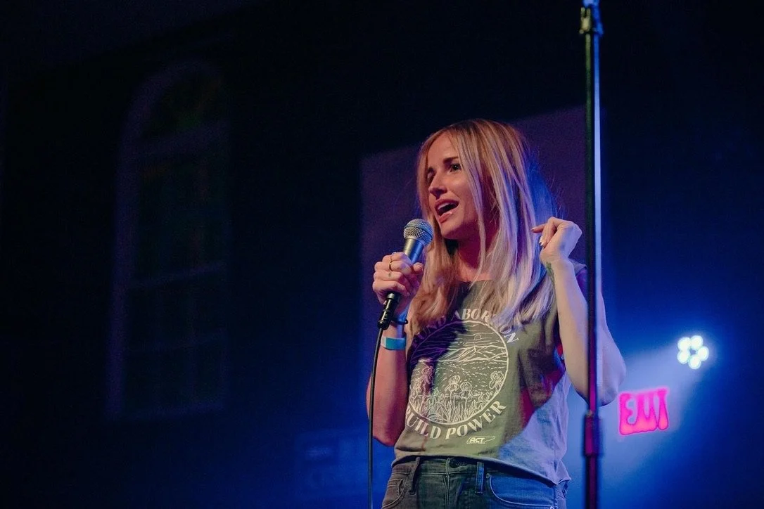 A young woman with long blonde hair singing or speaking into a microphone on stage with colorful lighting.
