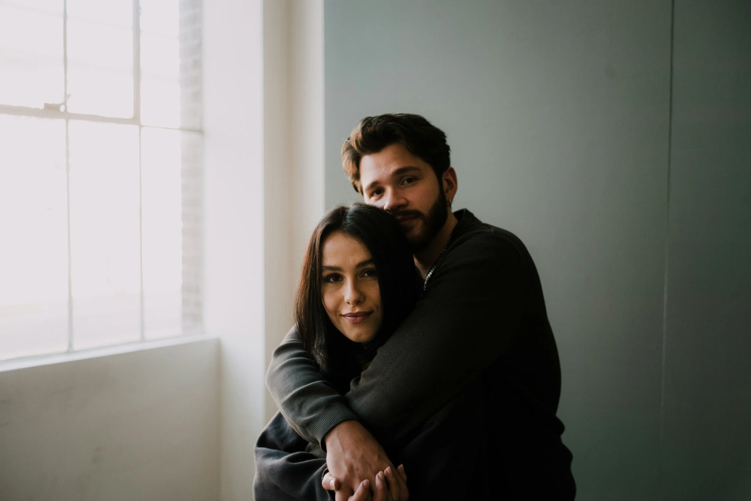 A young man with dark hair and beard hugging a young woman with long dark hair indoors near a window.