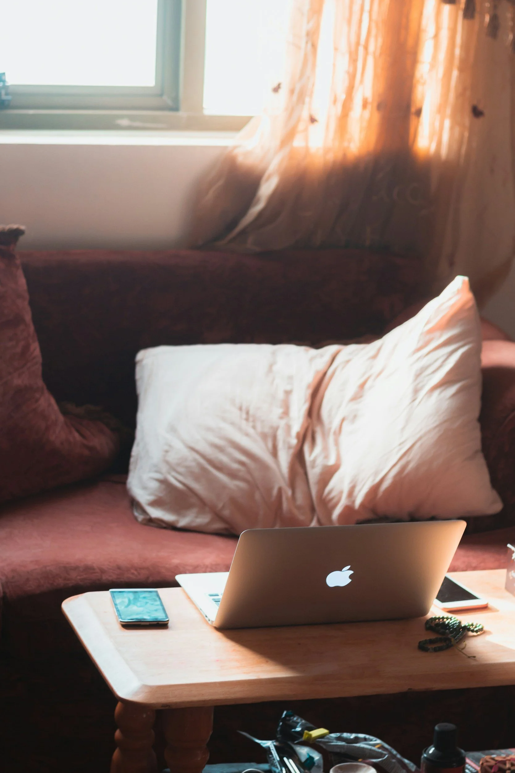 A living room with a window, curtains, a sofa with pillows, a wooden coffee table with a laptop, smartphone, and other items, and an open laptop on the table.