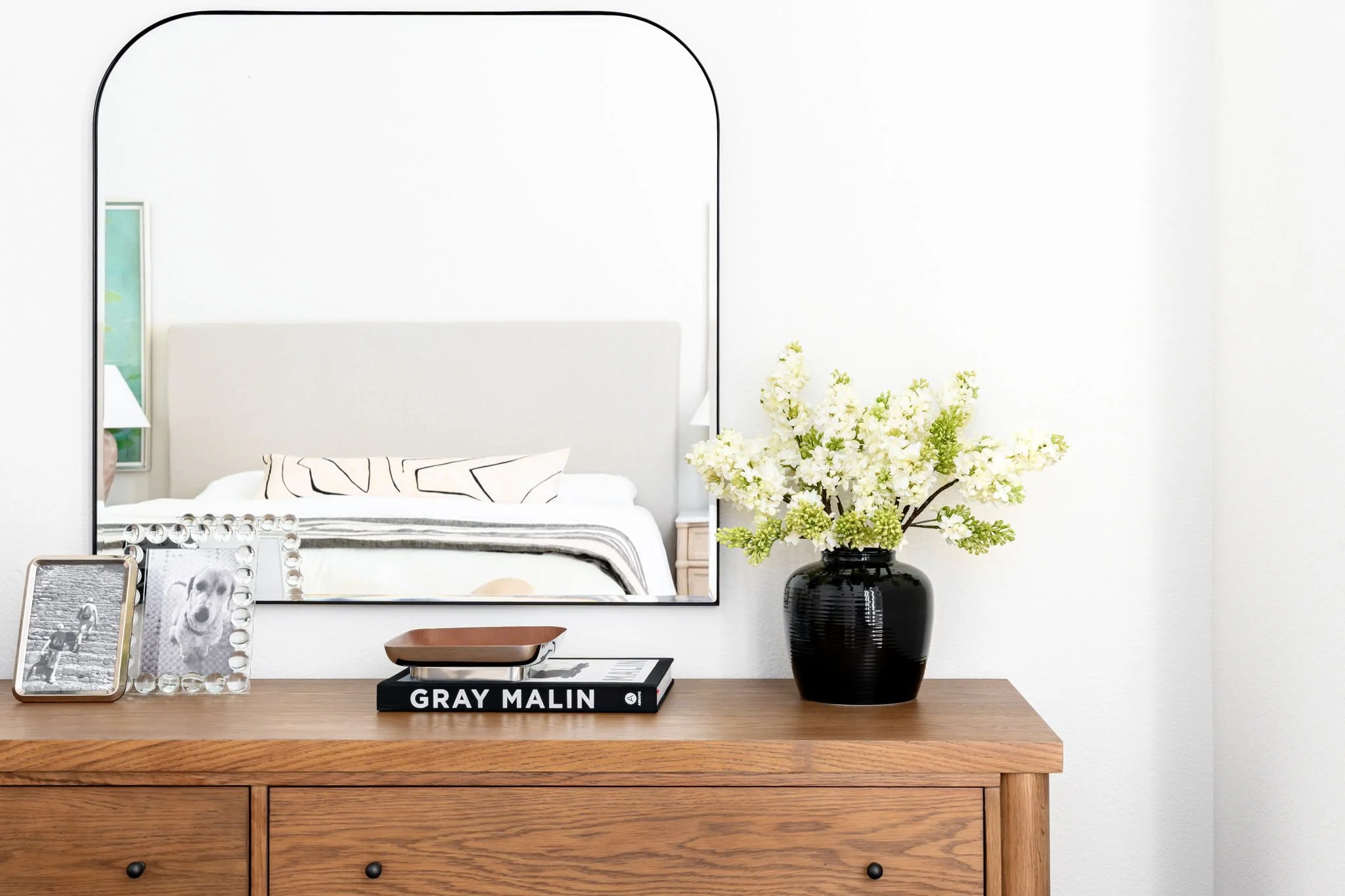 wood dresser with black vase of white flowers and arched mirror hung above. reflection of linen upholstered bed seen in mirror