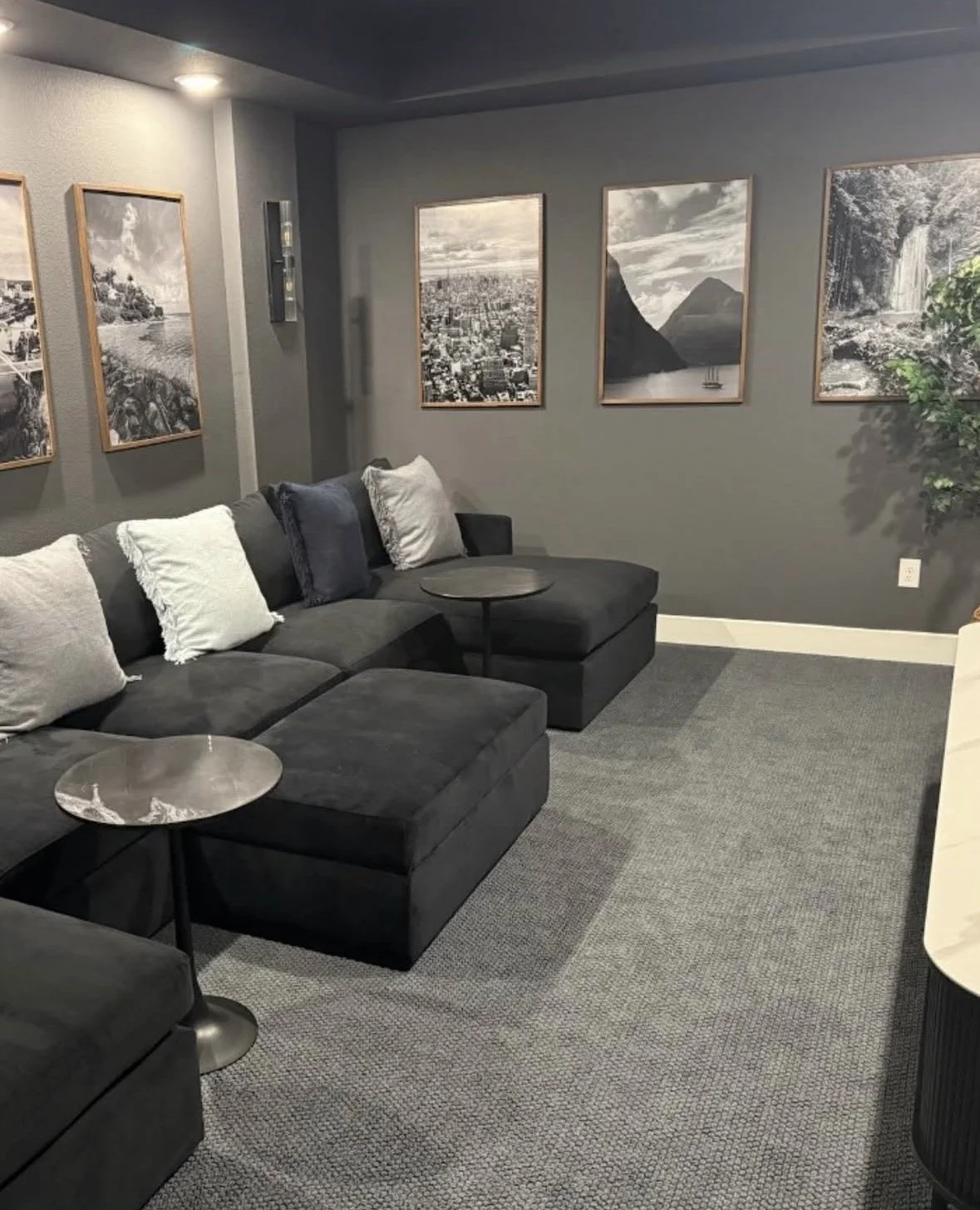 A media room in Prosper, Texas after an interior design service from Brooke Bundy Designs. The room features cozy low lighting, framed photos on the walls of travel destinations, a long couch with foot rests, and pillows and small side tables.