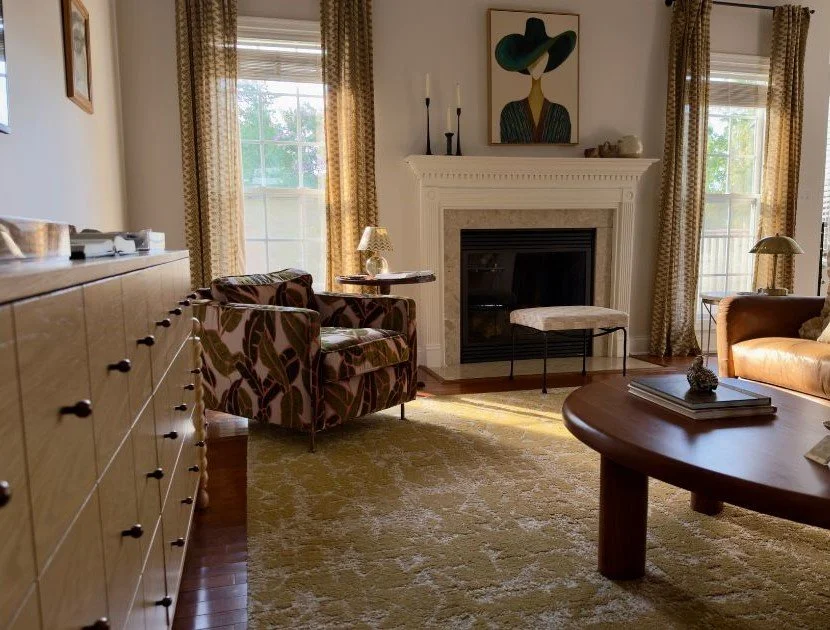 living room with wood dresser, green and pink patterned chair, wood coffee table and yellow rug. A green and yellow portrait is above the fireplace. 