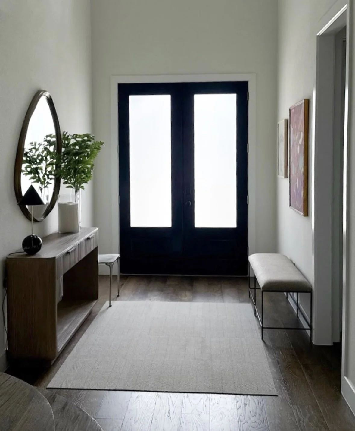 An entryway in Prosper, Texas, after interior design services by Brooke Bundy Designs. Photo shows a console table with a tear-drop-shaped mirror over it, a faux plant, a candle, a small lamp, a chair, and a rug.