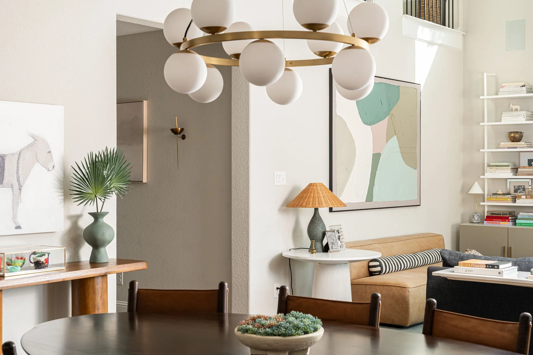 dining room with dark wood table, blue and red flowers on table, gold chandelier. living room visible with leather sofa and blue and green abstract art.