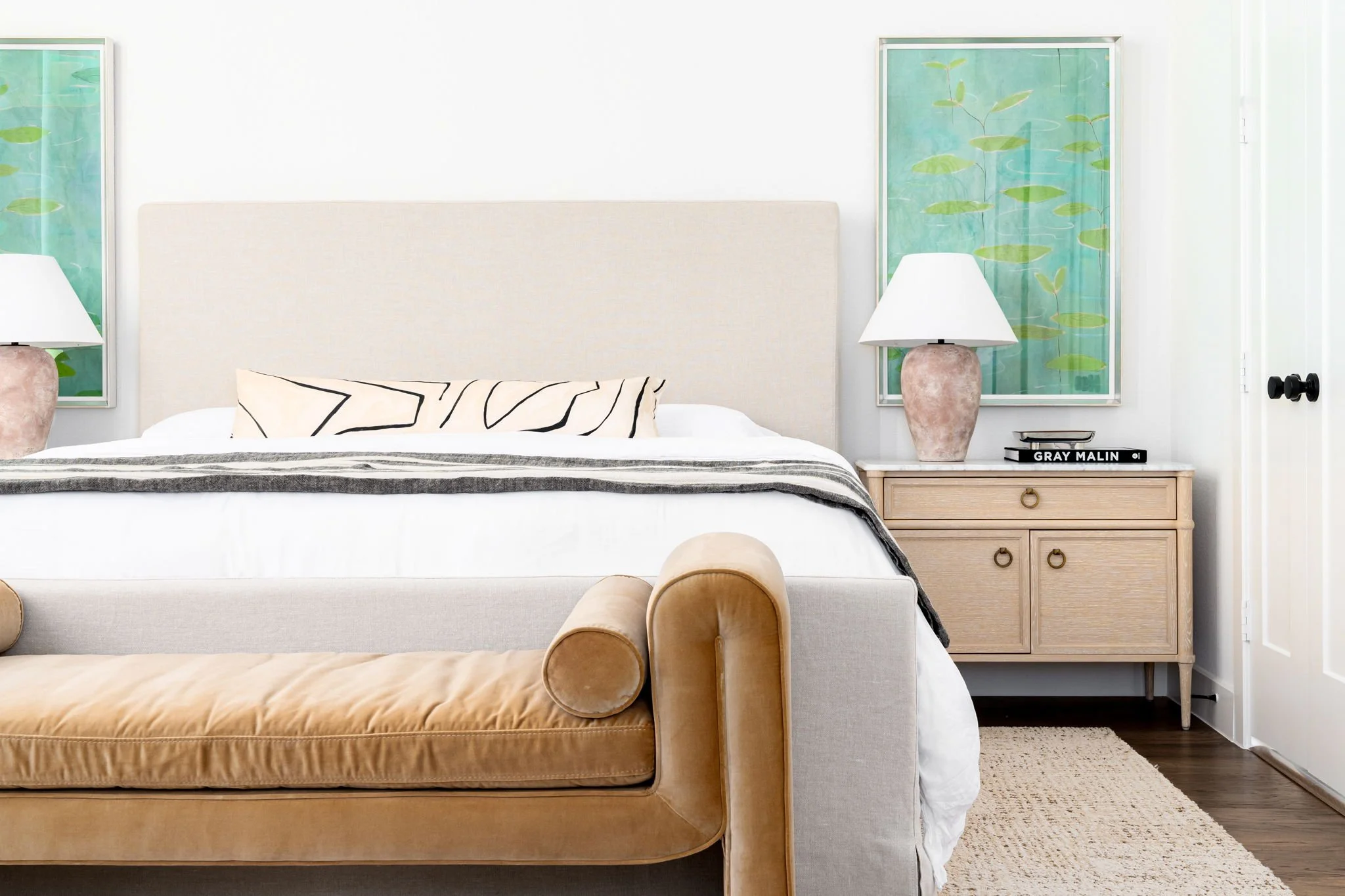 primary bedroom with linen upholstered bed, gold upholstered bench, light wood nightstands, terricotta lamps and green wall art