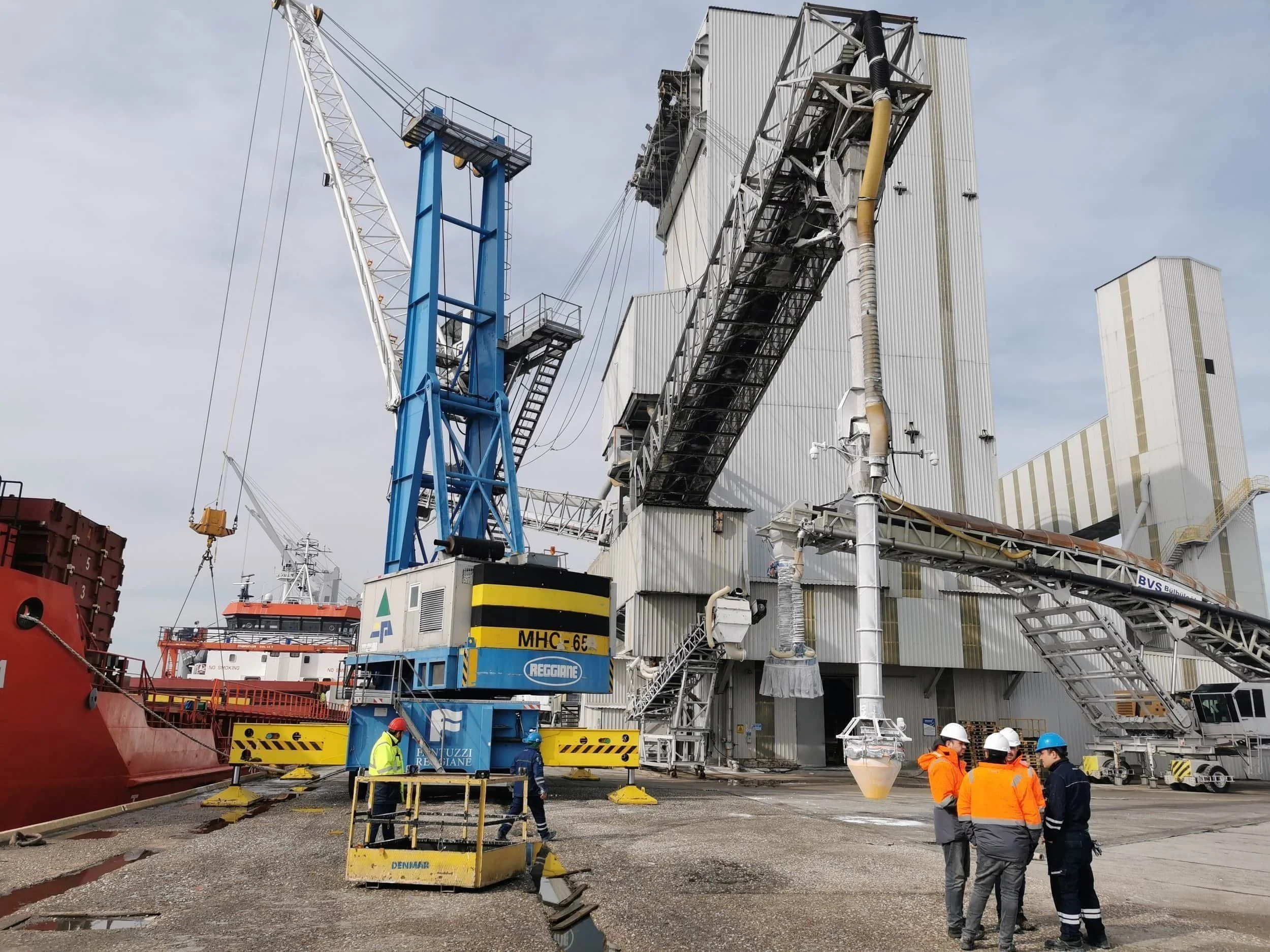 Large dust suppression hopper suspended from a chute on a structure extending from a storage building at a bulk loading area. A group of people in hi-vis and hardhats stand nearby. Cranes and a ship are visible in the background
