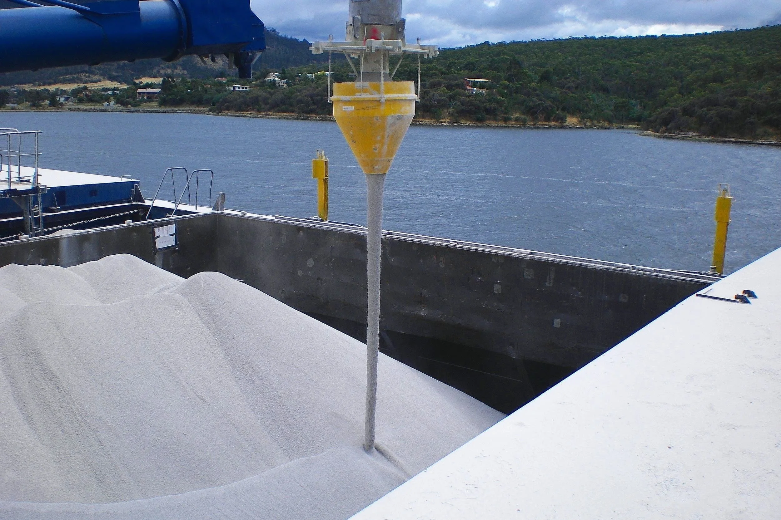 A white granular material, is in the process of being transferred from a ship or dock using a dust suppression hopper, with water, trees, and hills in the background.