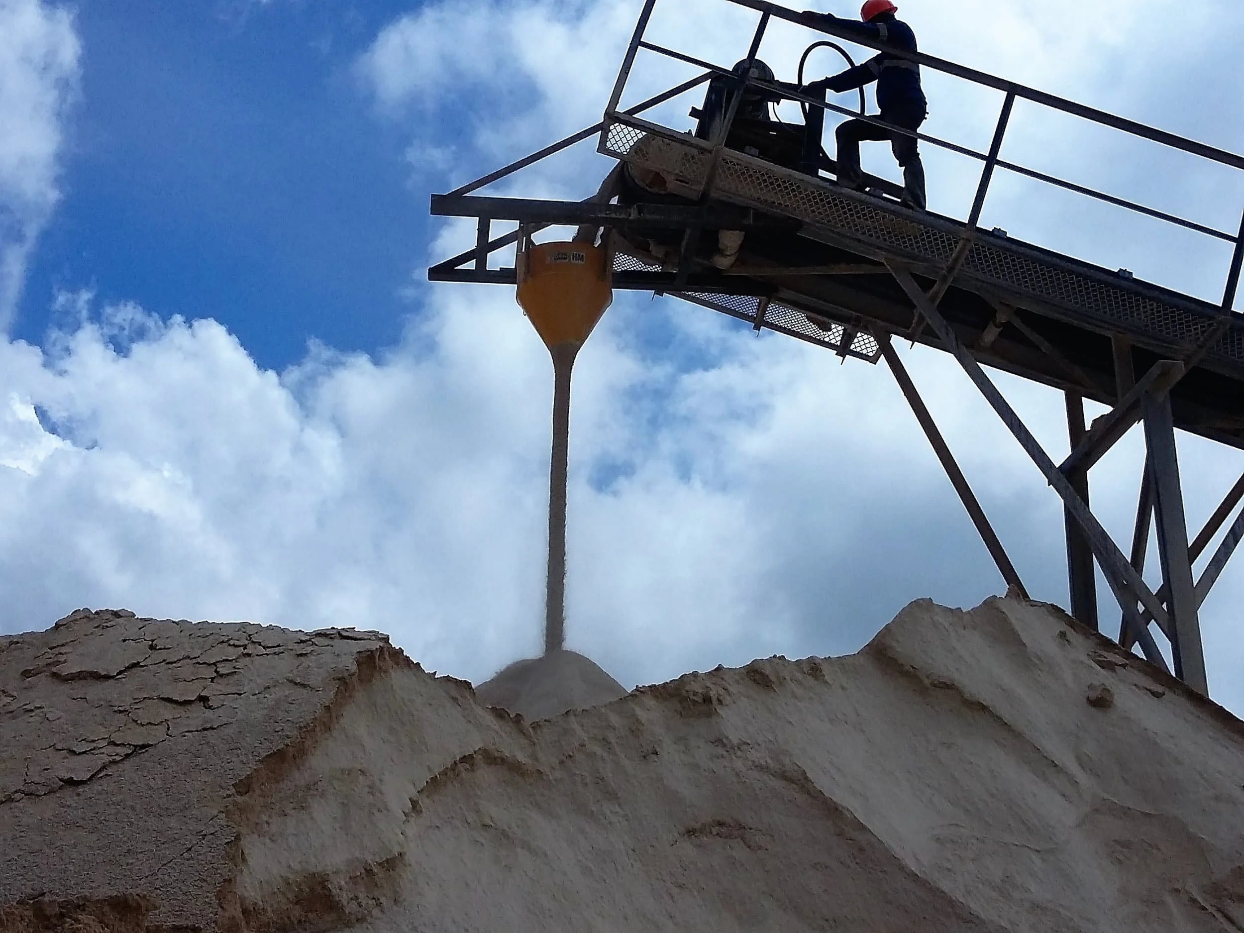 Two workers on a tall industrial platform pouring a light-colored powder or mineral into a pile below, using a dust suppression hopper with partly cloudy sky in the background.