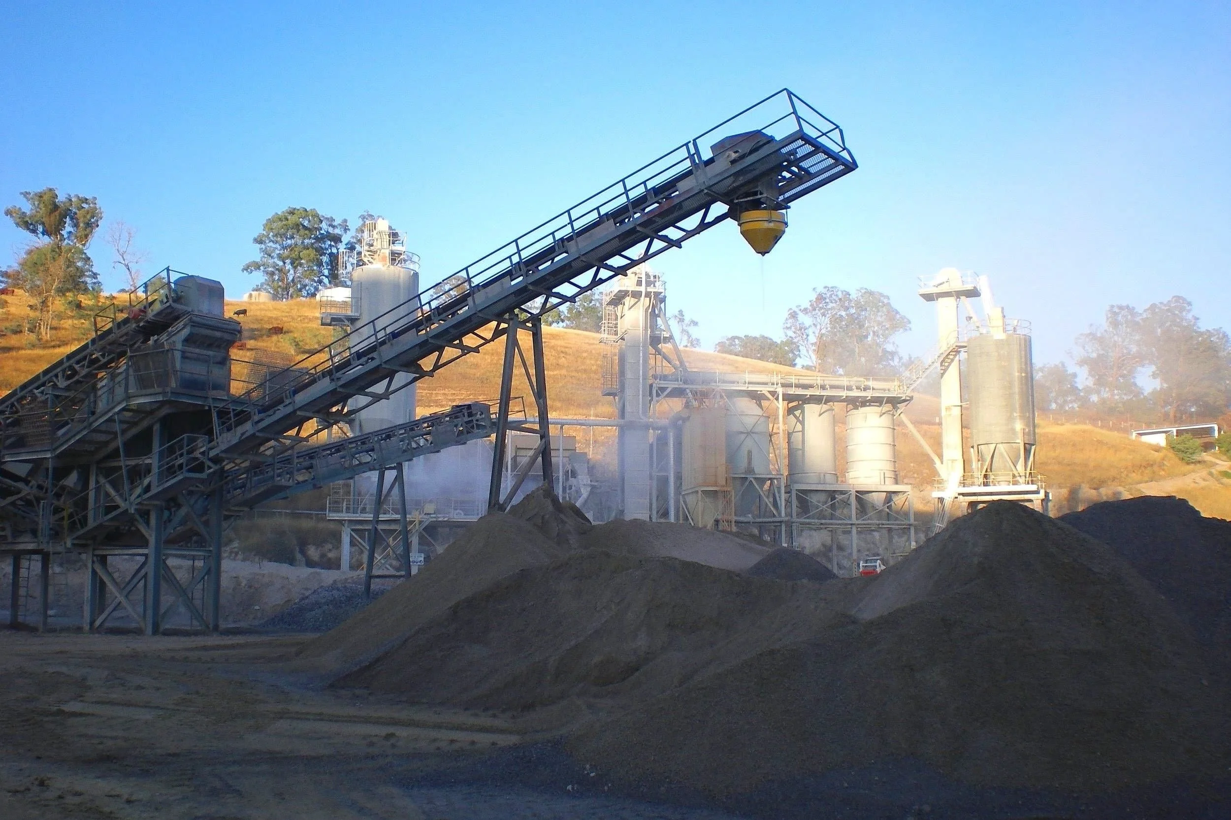 An industrial mining site with large metal structures, conveyor belts, a dust suppression hopper and piles of dirt and gravel in the foreground.