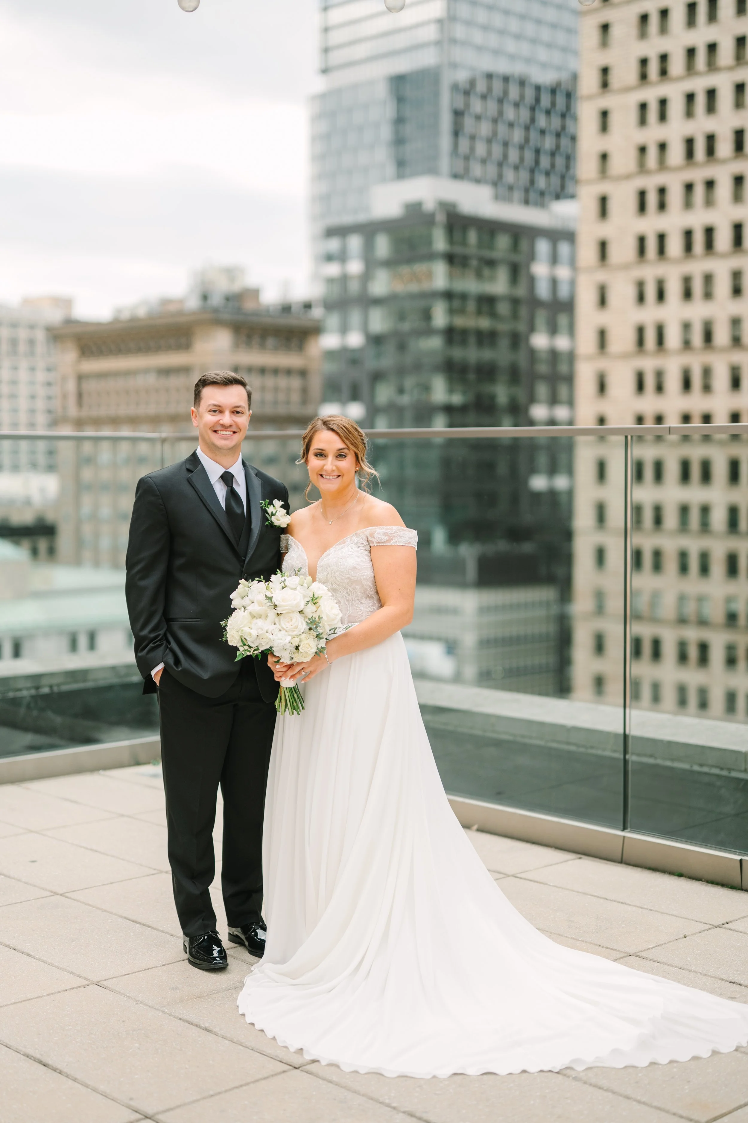 A smiling bride and groom, standing on the rooftop of Hotel Monaco, with the Pittsburgh city in the background.