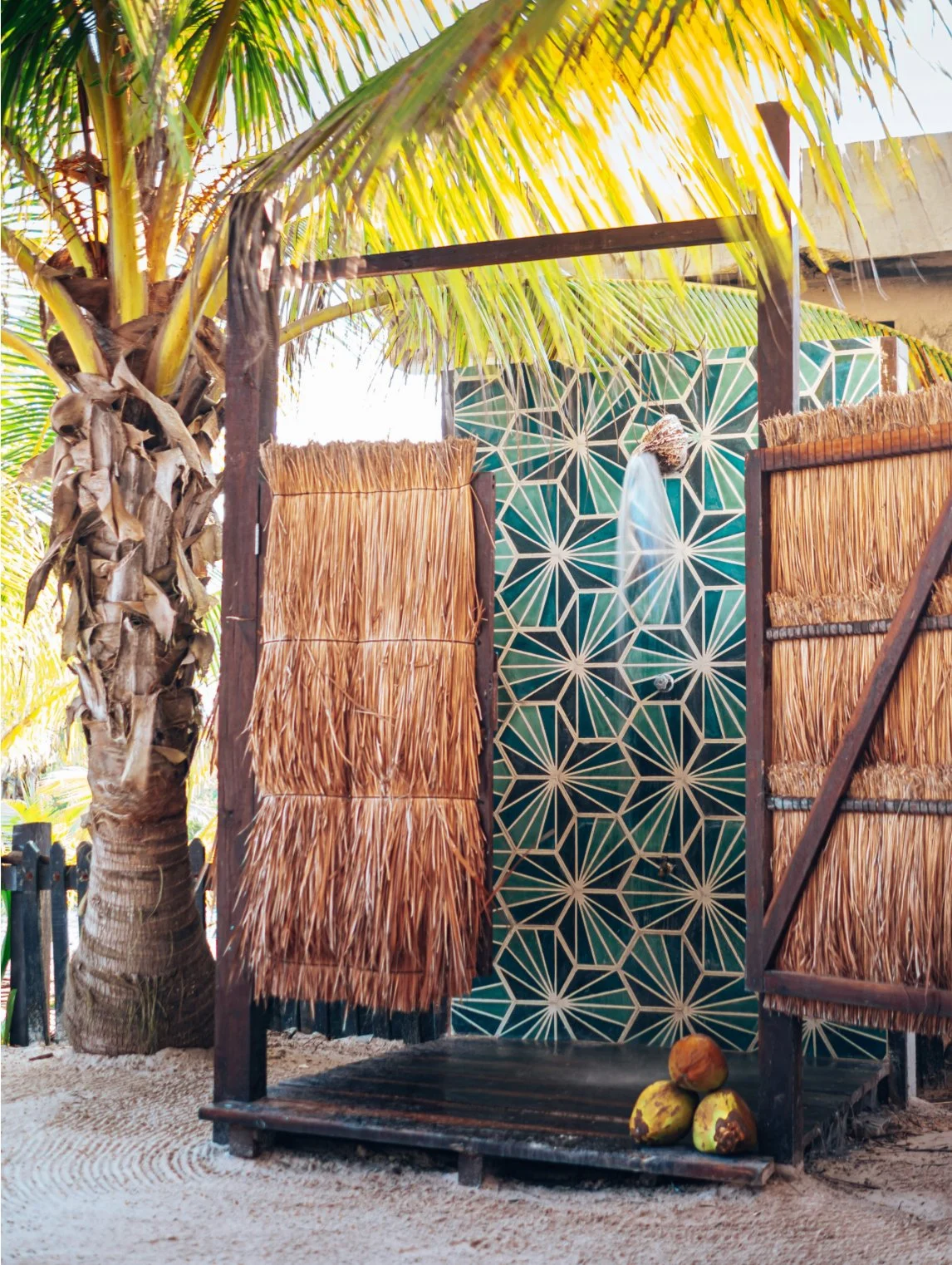 Outdoor shower on a sandy beach with palm trees, decorated with green tiles and hung thatch curtains, with coconuts on the ground.