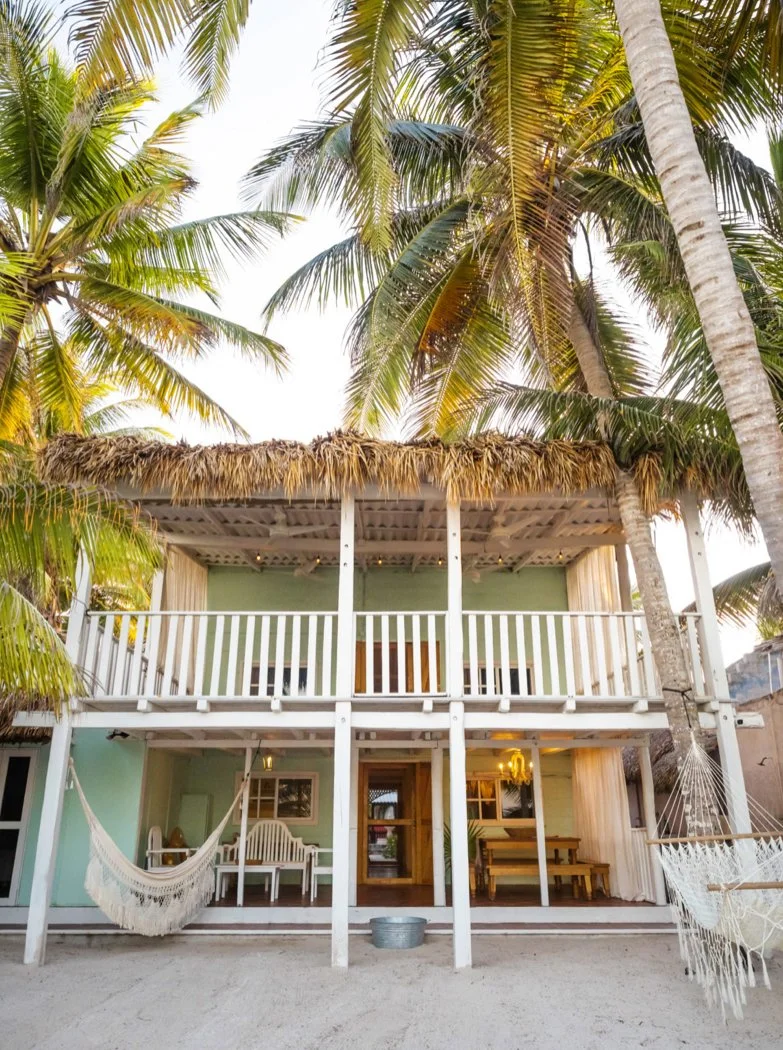 A two-story beach house painted in light green with a thatched roof over the upper balcony, surrounded by tall palm trees, with hammocks hanging in the front on sandy ground.