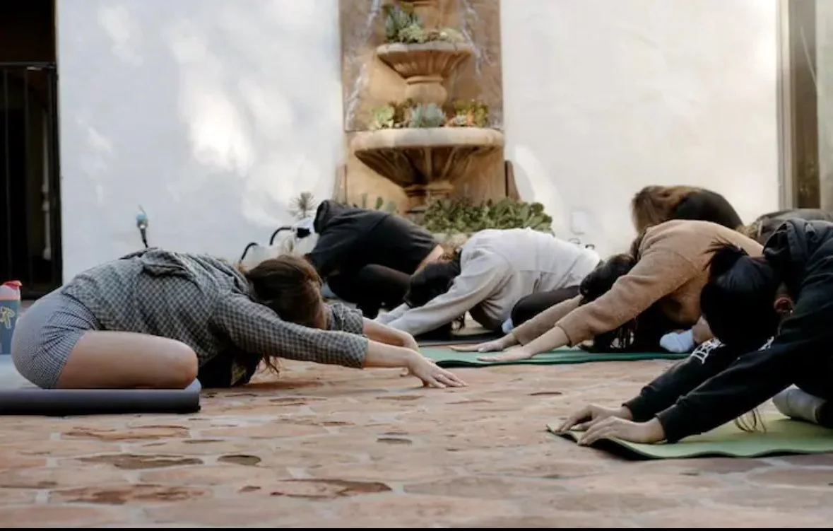 People participating in a yoga class in a spacious indoor setting, kneeling on yoga mats with arms extended forward.