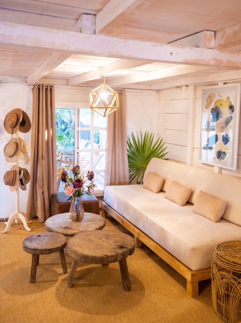 Cozy living room with white wooden walls and ceiling beams, beige curtains, a large window, a light fixture hanging from the ceiling, a white sofa with beige pillows, a large green plant, a small round wooden table with a vase of flowers, a hat stand with hats, and a framed abstract artwork on the wall.