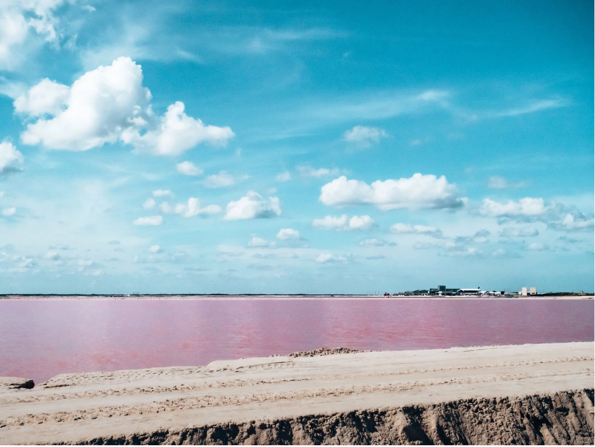 Pink salt lake with a sandy shoreline and a blue sky with clouds in the background.