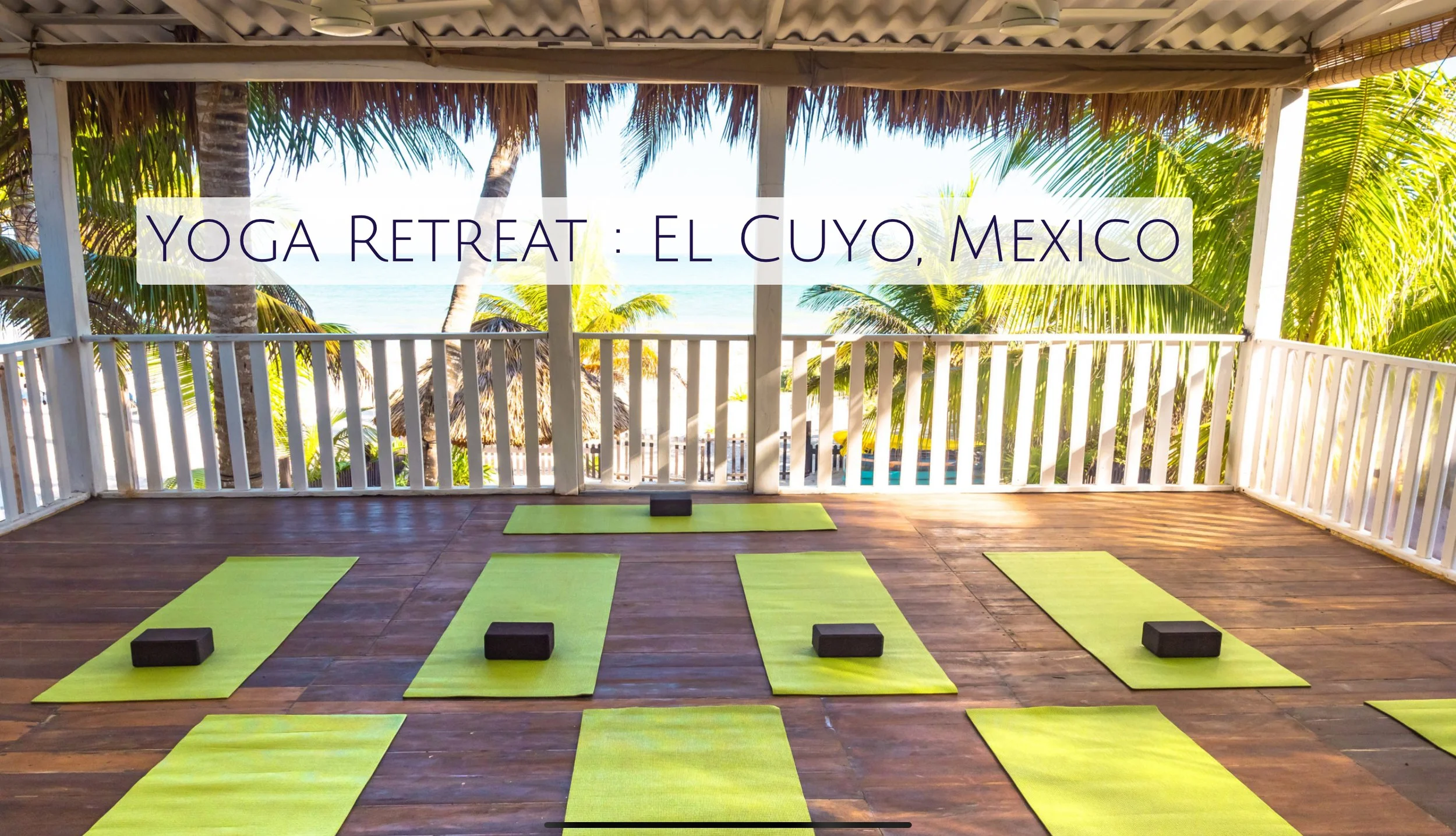 Yoga mats and blocks set up on a wooden deck with a view of palm trees, ocean, and beach in the background during a sunny day at a beachside yoga retreat in El Cuyo, Mexico.